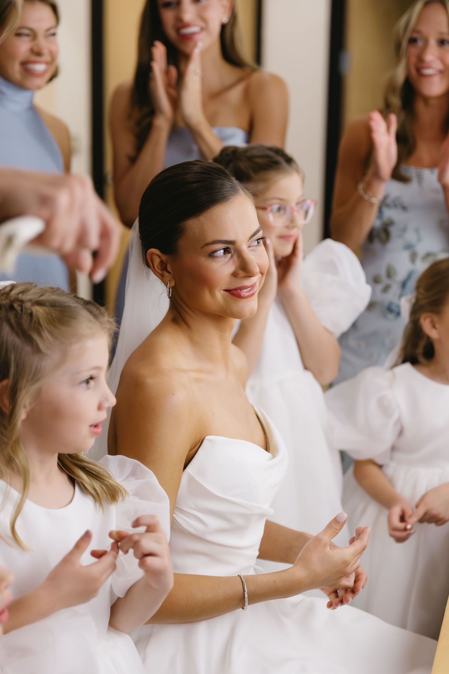 A wedding party gathering shows joyful expressions and white dresses in an intimate indoor setting.
