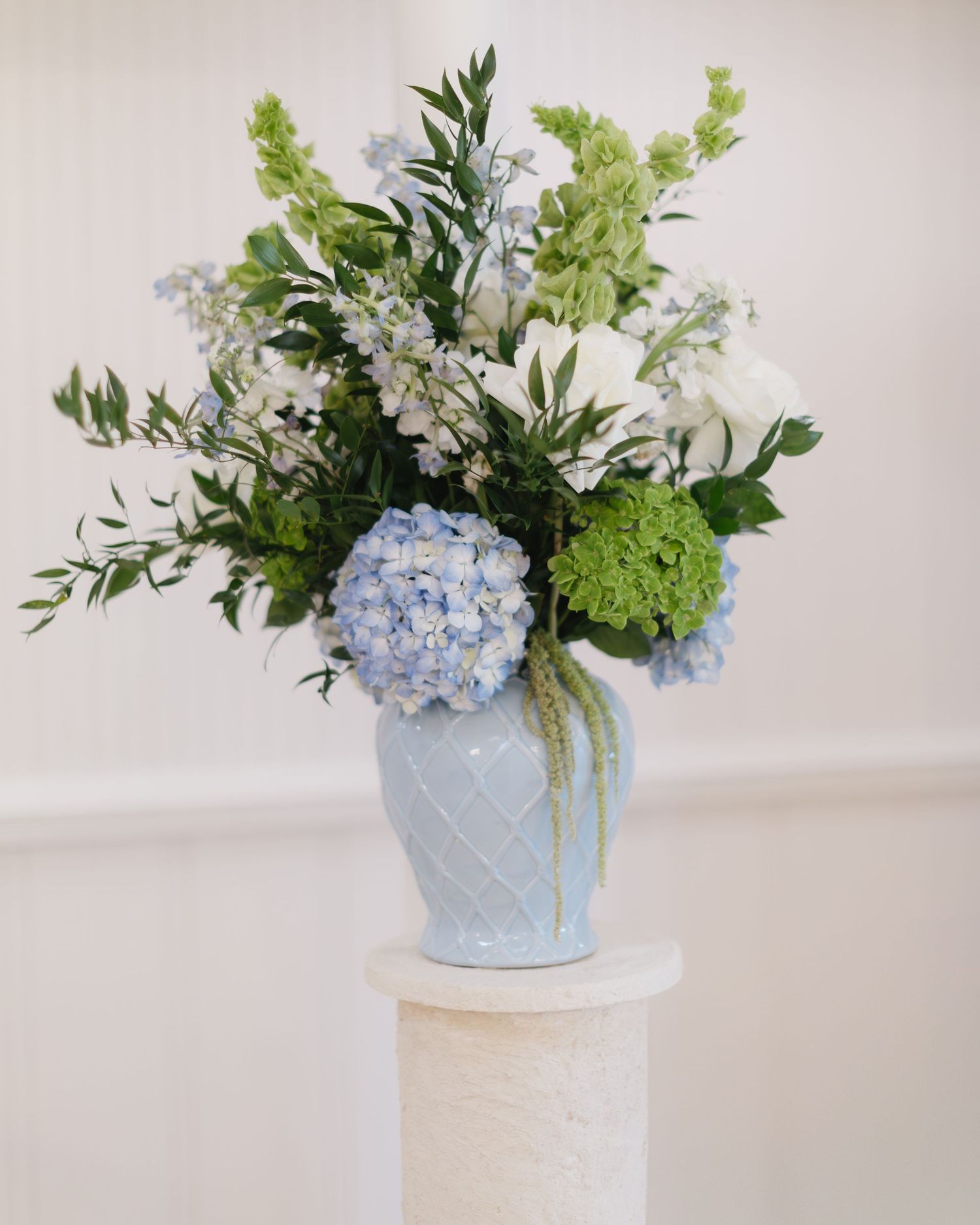 Blue hydrangeas and white flowers arranged in a light blue vase on white pedestal.