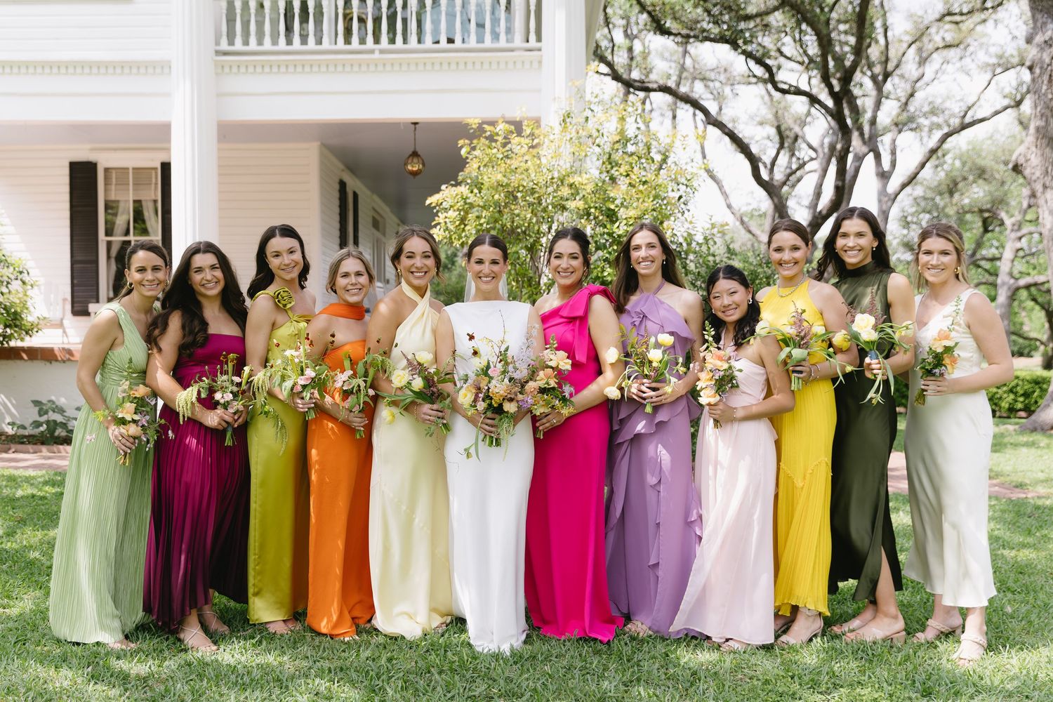 A group of bridesmaids stand together outdoors wearing colorful dresses in green, magenta, orange, yellow and purple shades.