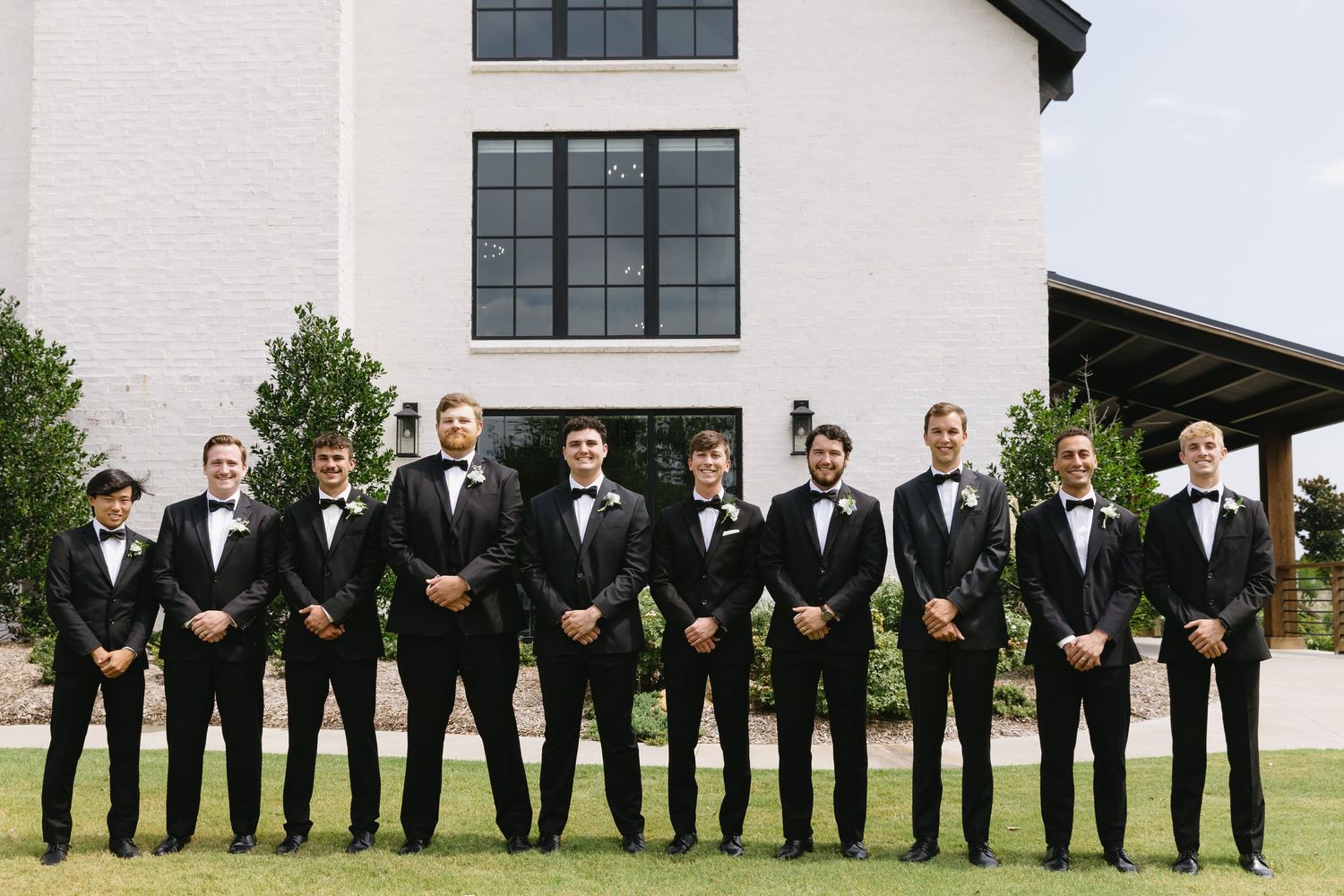 Groomsmen in black tuxedos pose in line outside white church building.