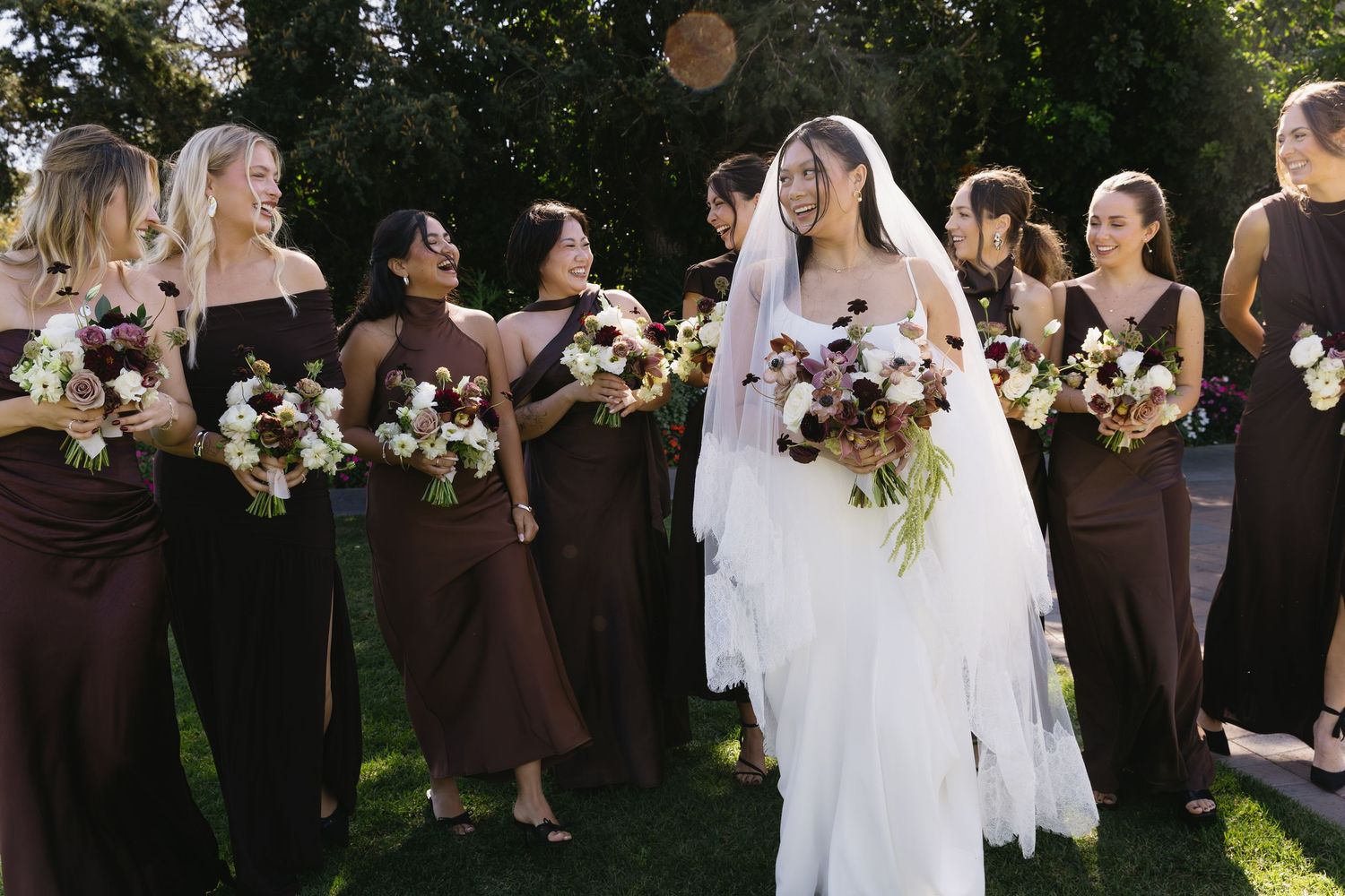 A bridal party in brown dresses poses with bouquets in an outdoor garden setting.