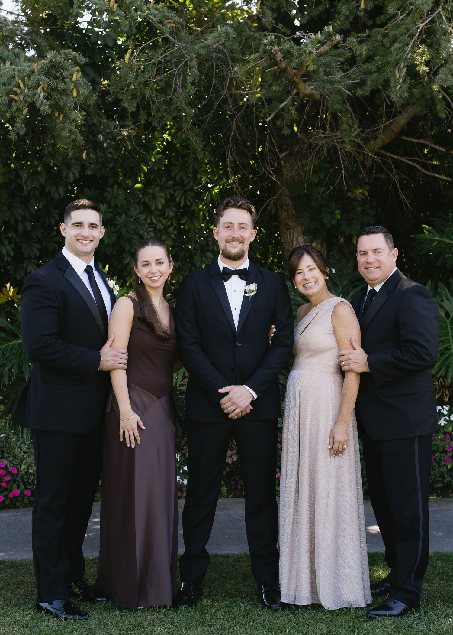 Wedding party group photo with members in formal attire against garden backdrop.