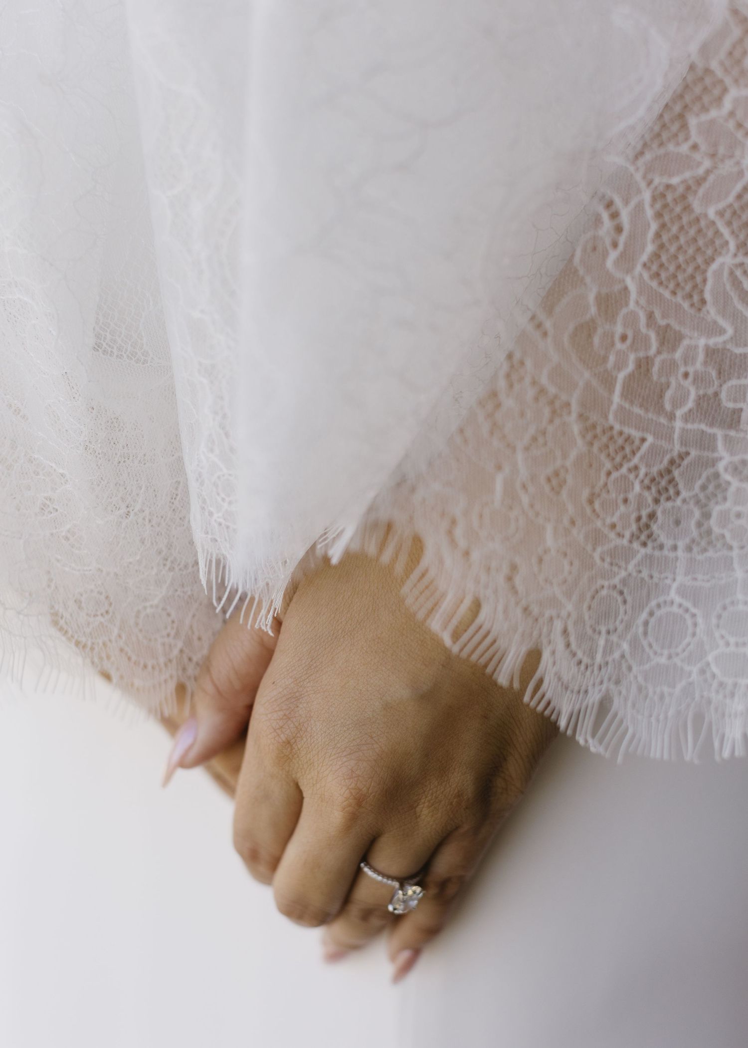 Close-up of a hand wearing a delicate engagement ring against white lace fabric.