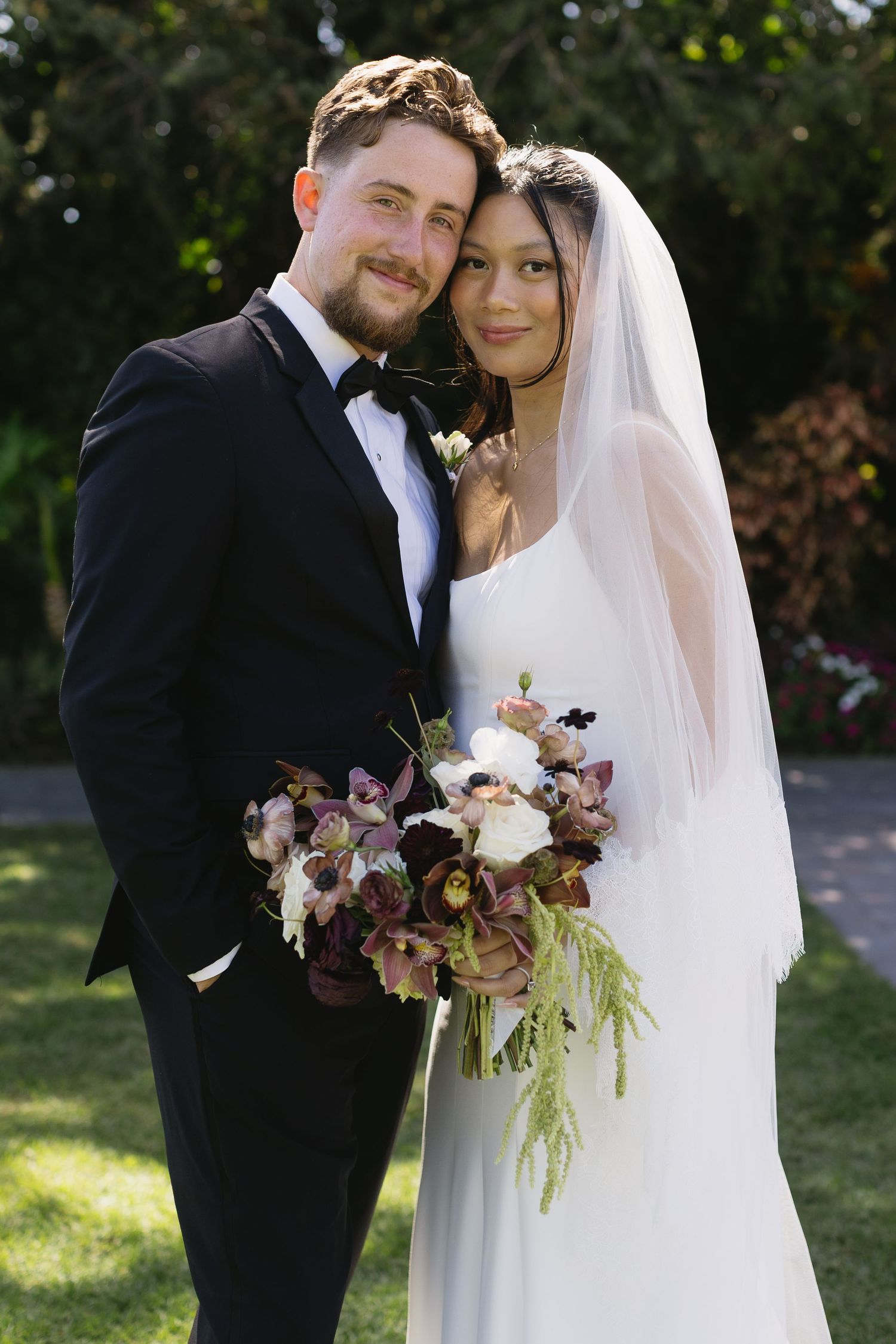 A wedding couple embraces outdoors with a beautiful floral bouquet.