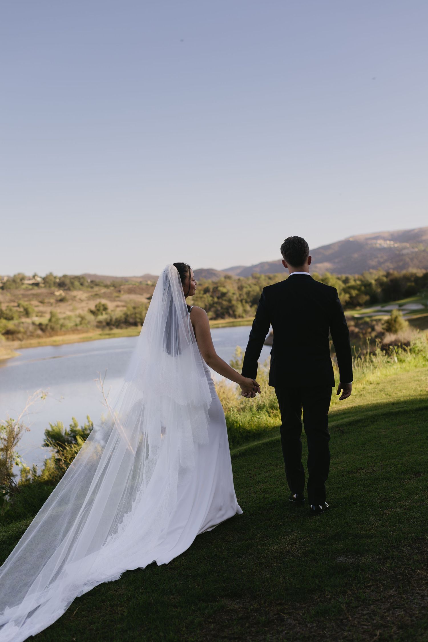 Bride and groom walking along grassy hillside with lake view in background.