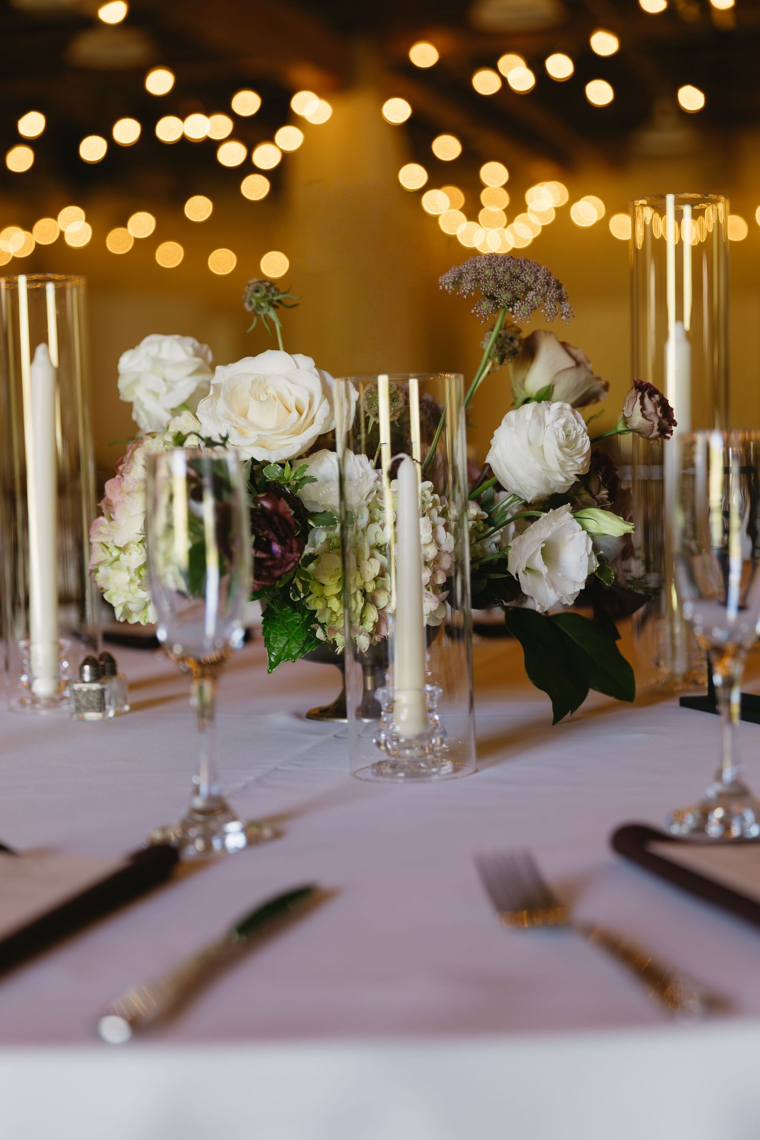 Elegant table settings with white roses and tall glass candleholders under warm reception lighting.