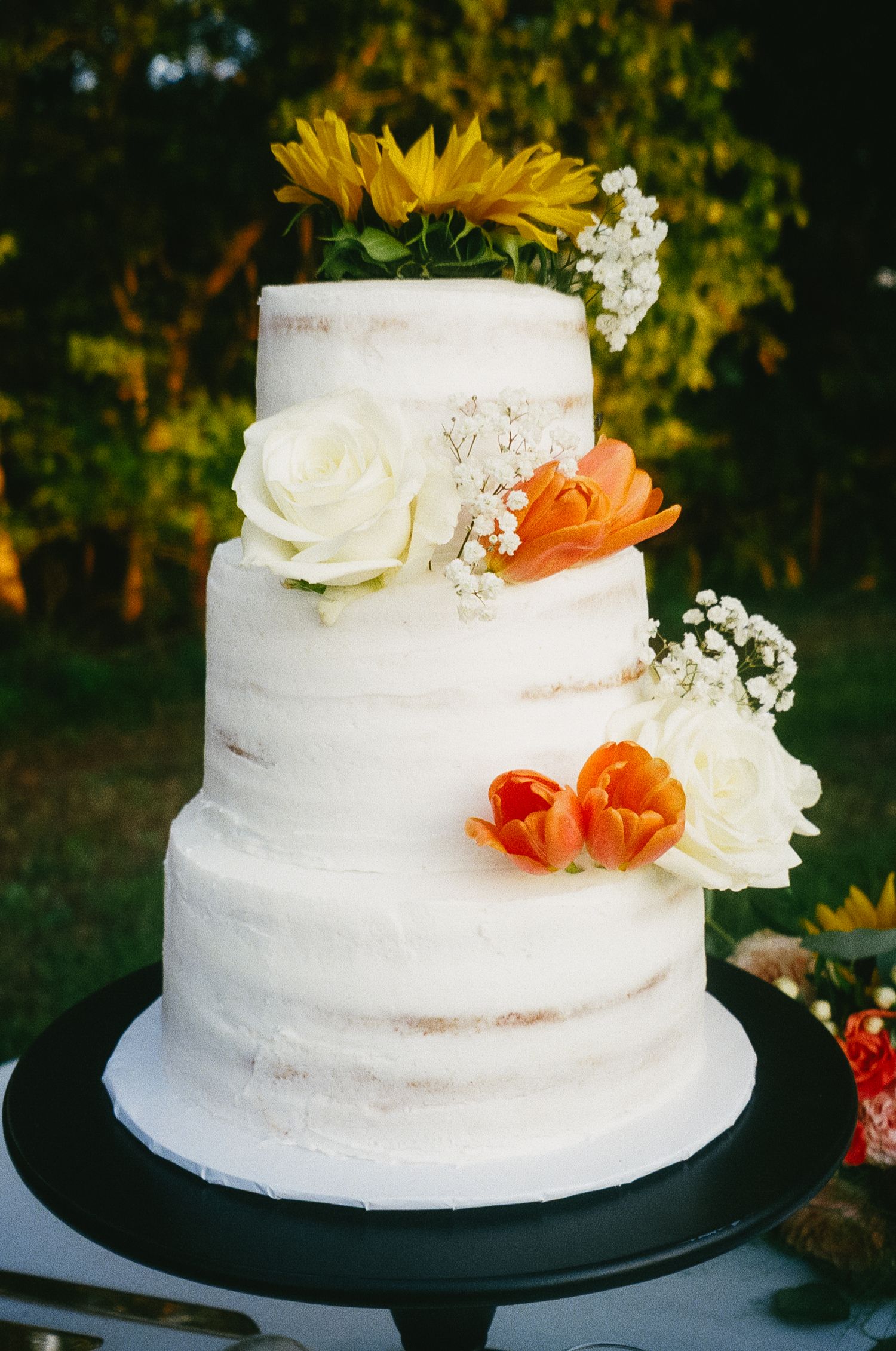 A white two-tiered wedding cake decorated with fresh orange and white flowers and baby's breath on a black cake stand.