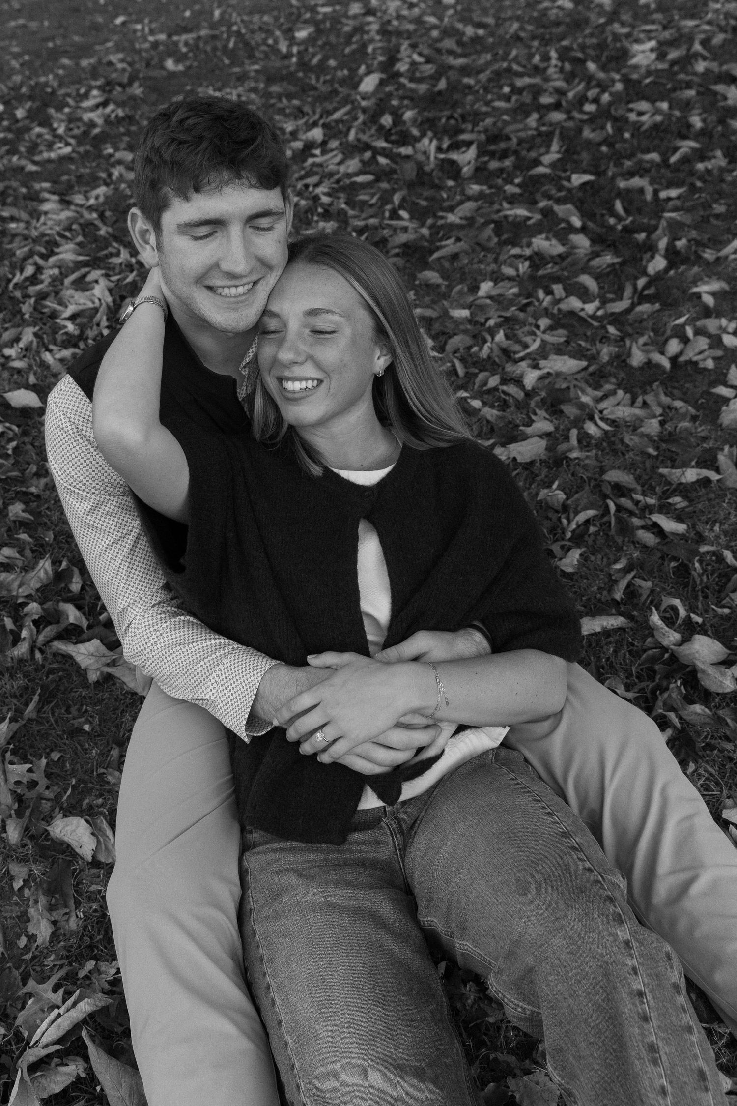 A couple shares playful moments while lying in fallen autumn leaves during a black and white photoshoot.