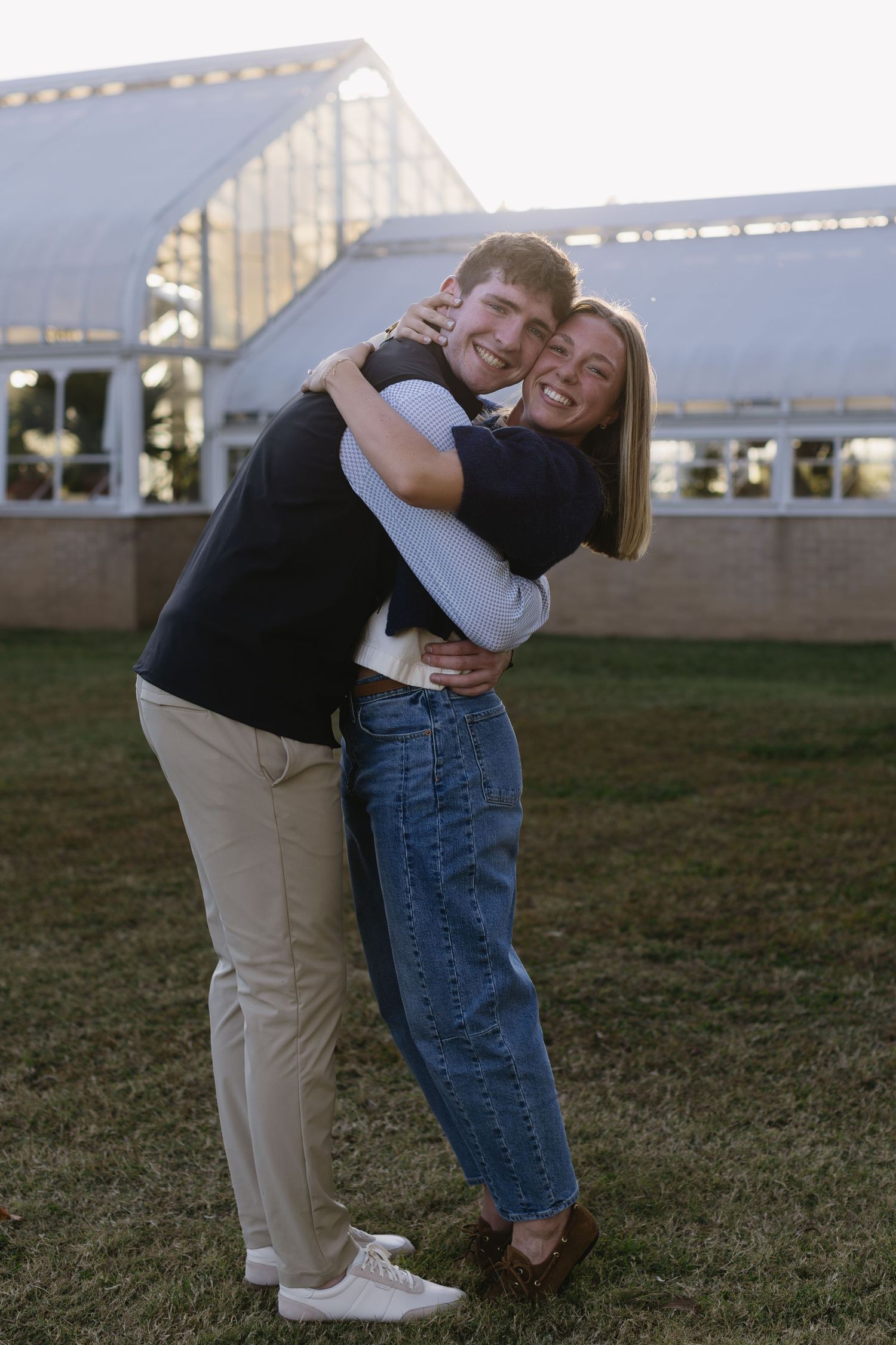A couple shares a joyful embrace while standing on grass with a greenhouse structure visible in the background.