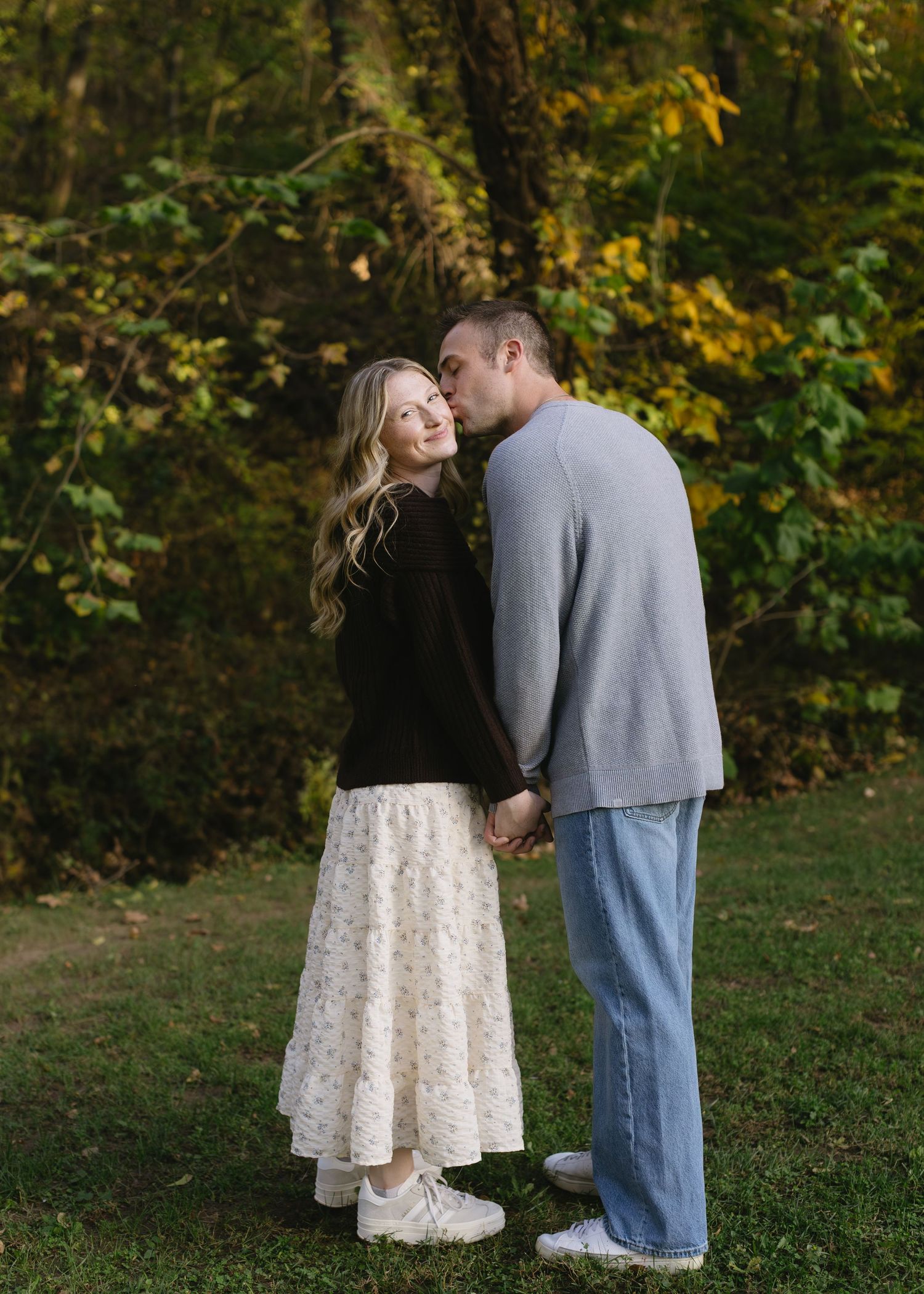 A couple shares a tender moment in an autumn park setting with colorful fall foliage in the background.