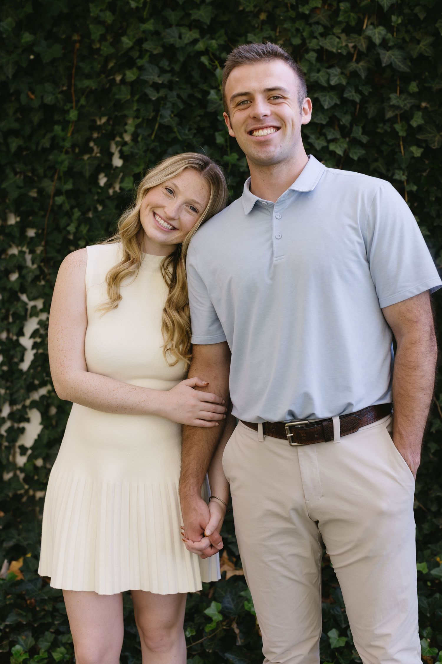 Couple in light summer outfits posing playfully against green foliage background.