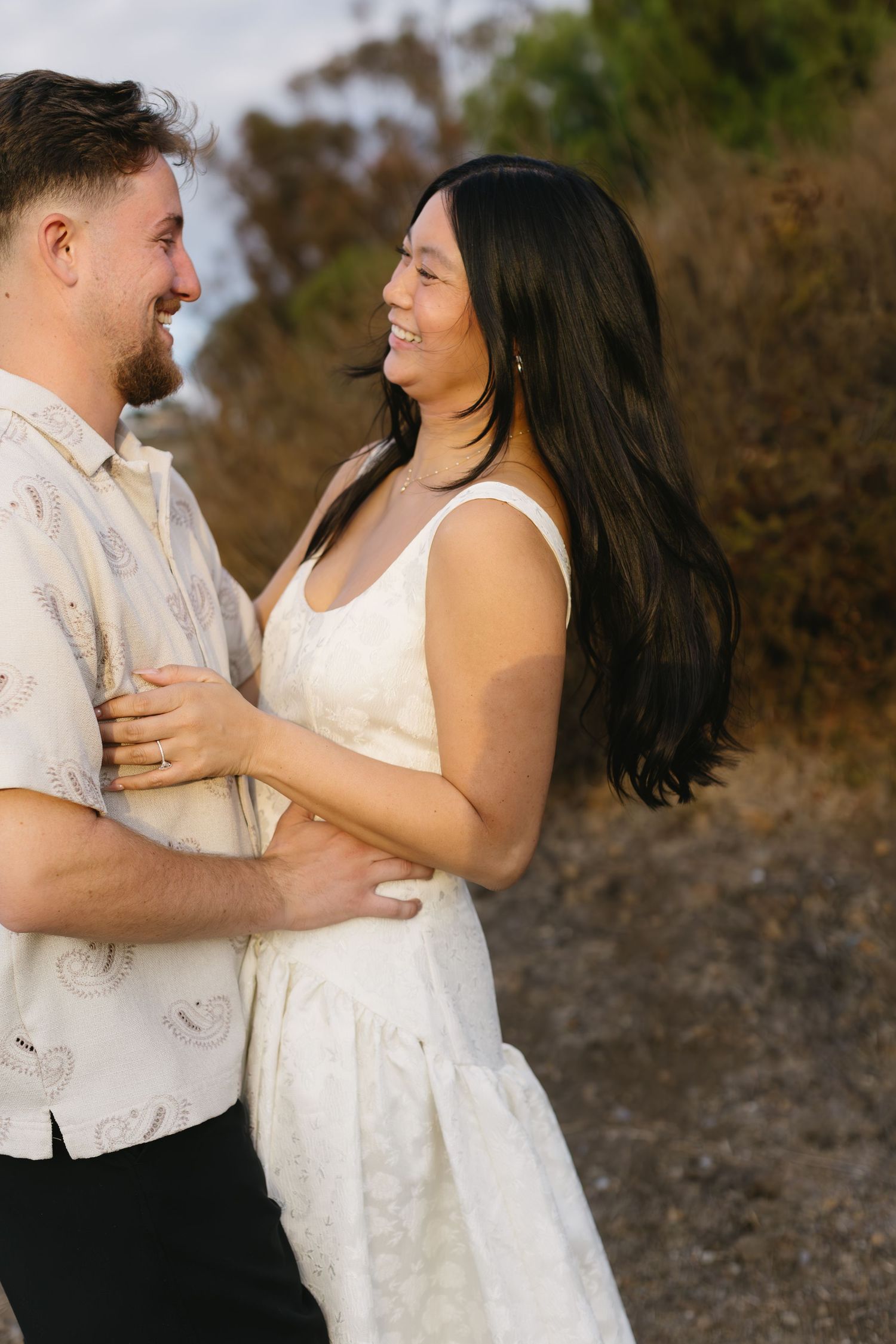 A couple embraces and laughs together during their outdoor photoshoot on a scenic hillside.