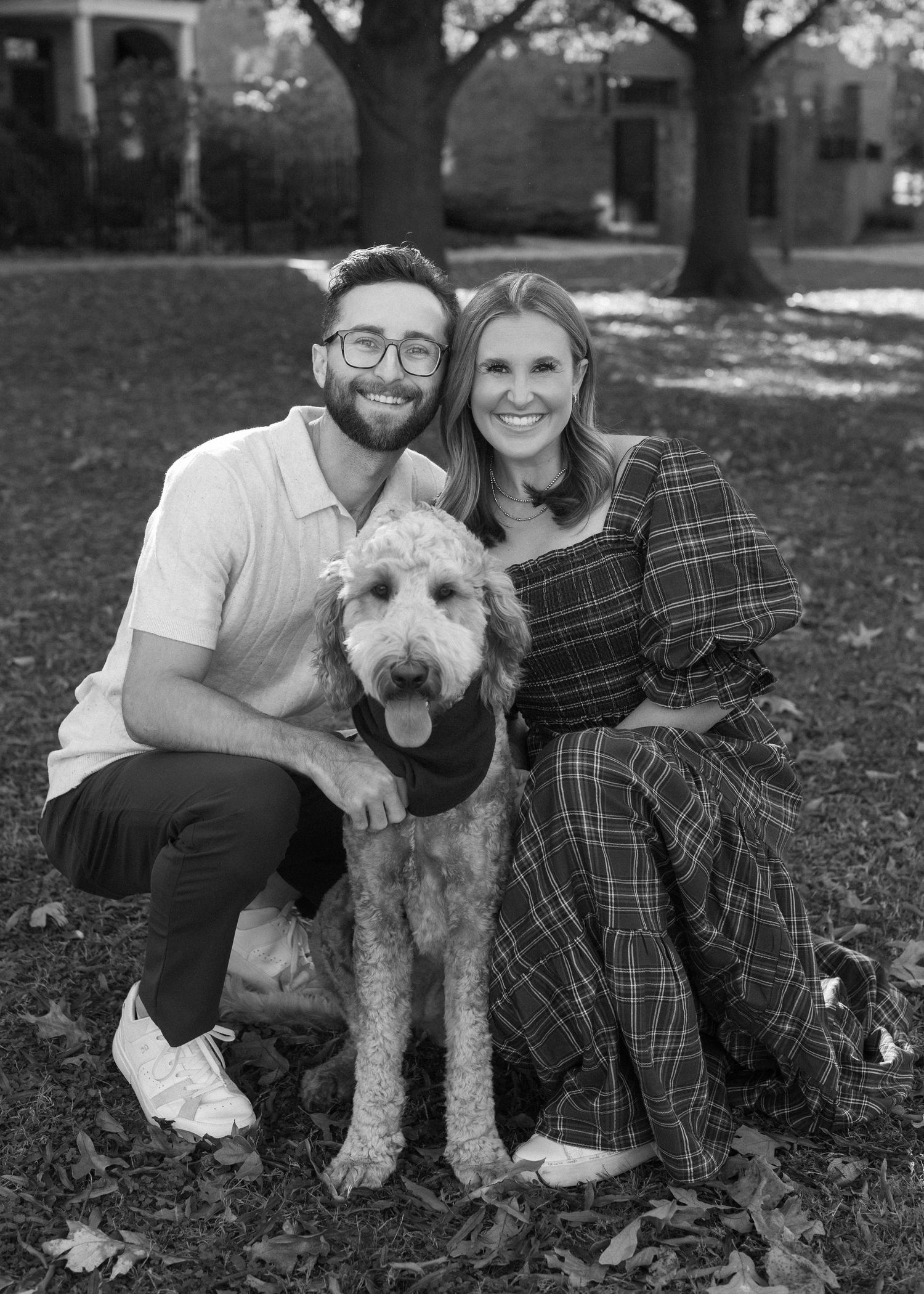 A black and white portrait of a couple with their Goldendoodle dog sitting among fallen leaves.