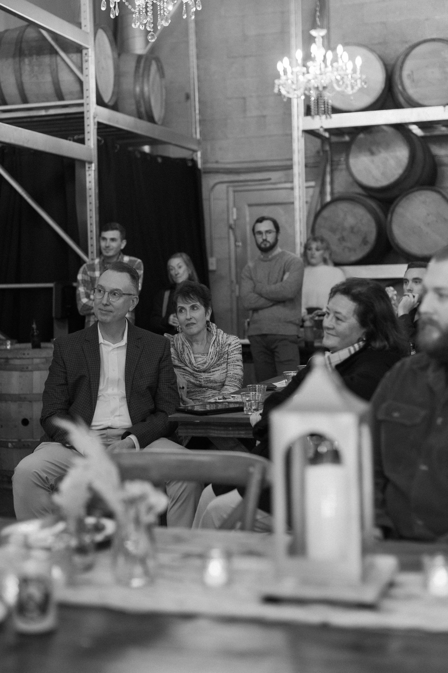 Guests seated in a rustic venue with wooden barrels and chandeliers in the background.