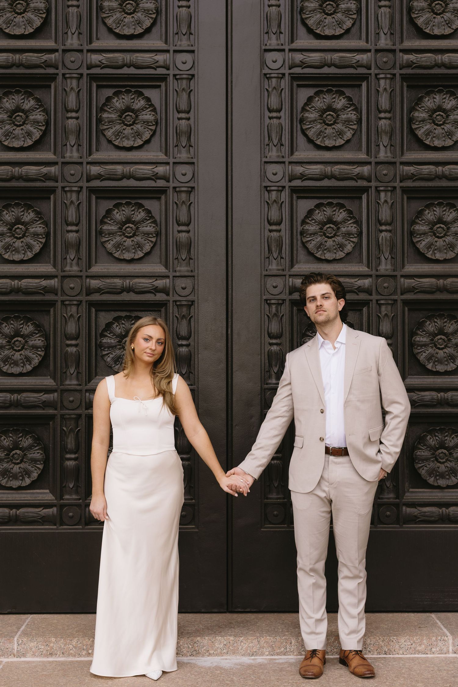 A couple in light colored formal wear poses playfully in front of a decorative black door.