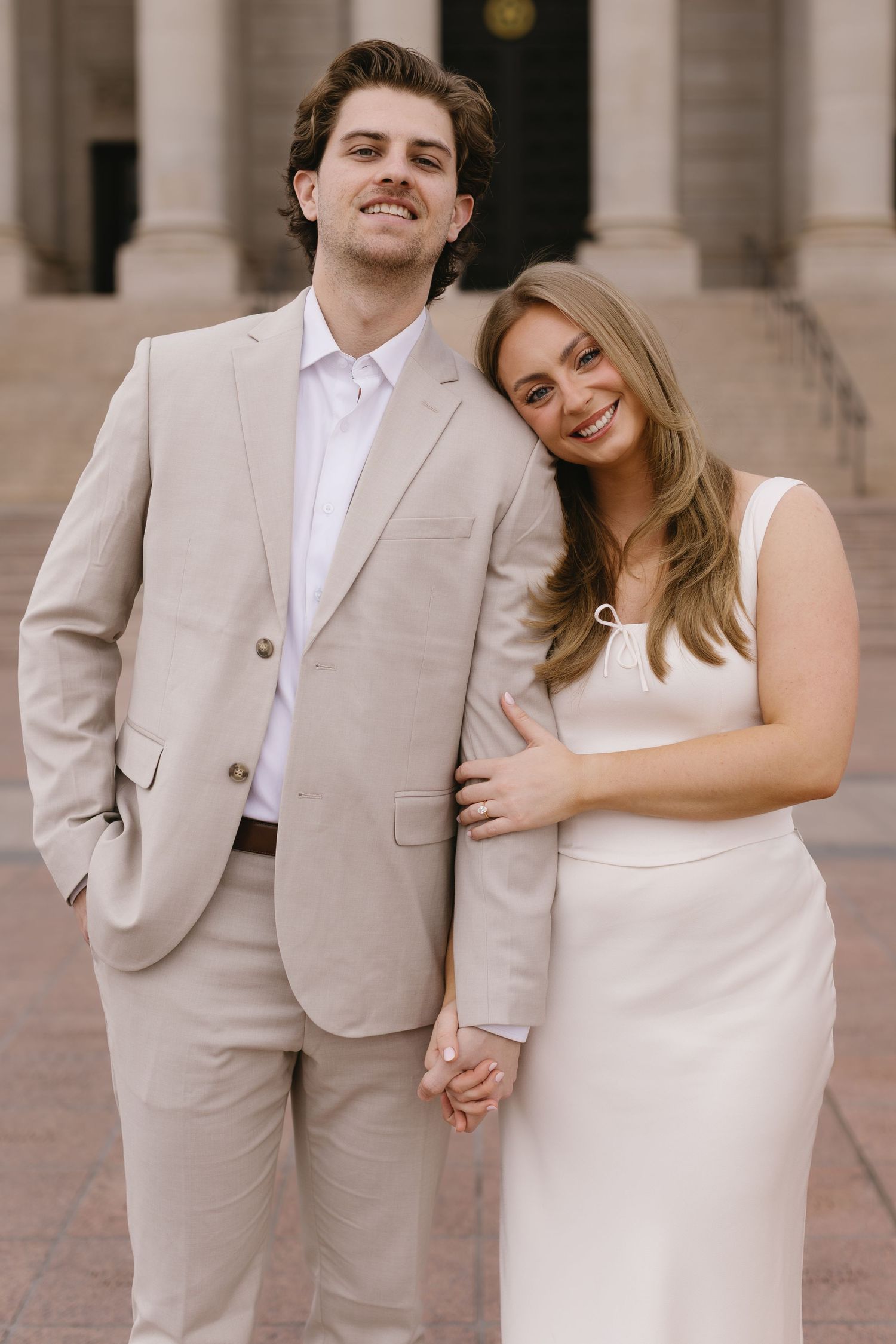 A couple in coordinated beige and white wedding attire embraces on stone steps of a grand building.