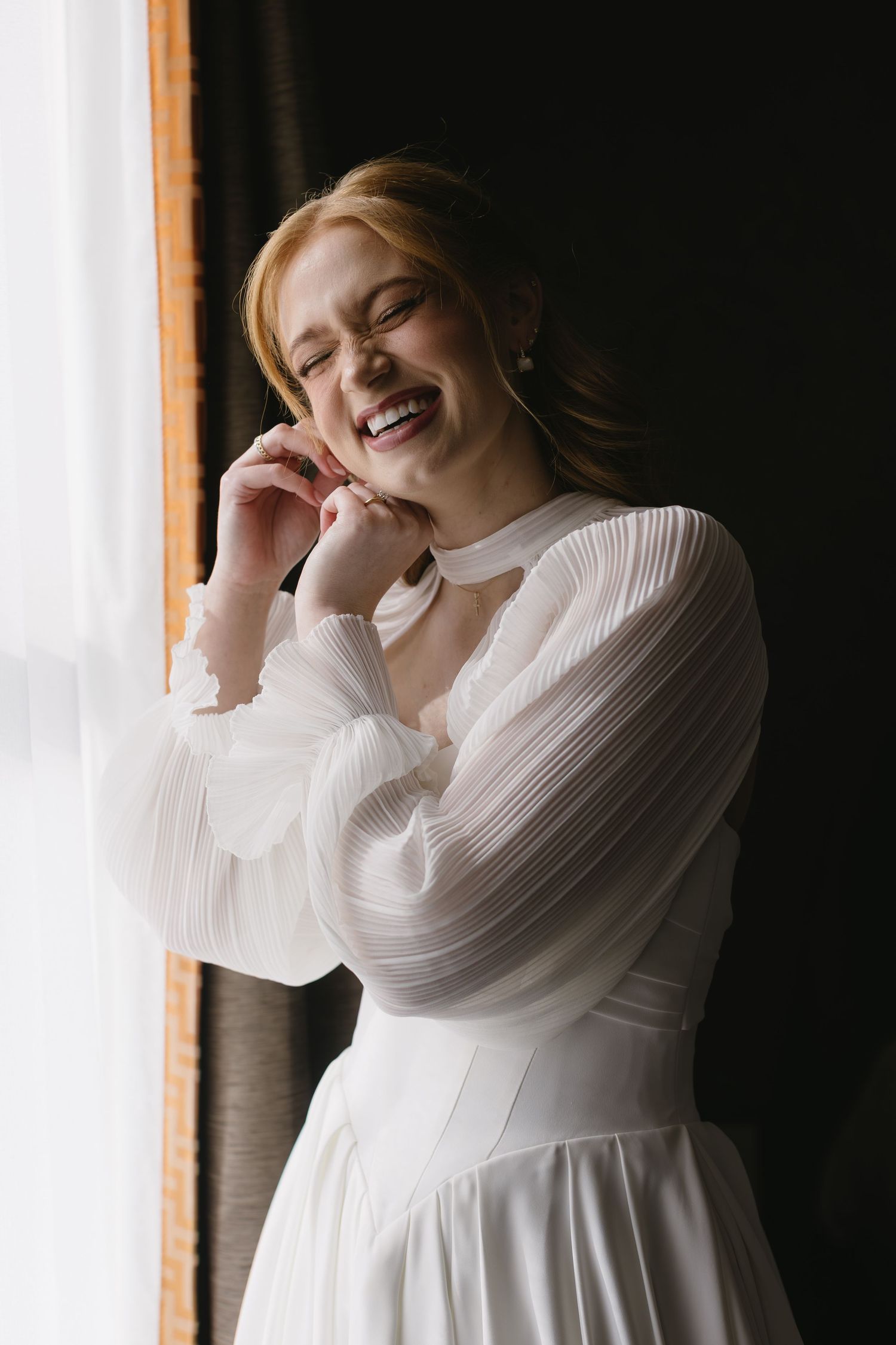 A bride in a flowing white dress poses elegantly by a window in romantic bridal portraits.