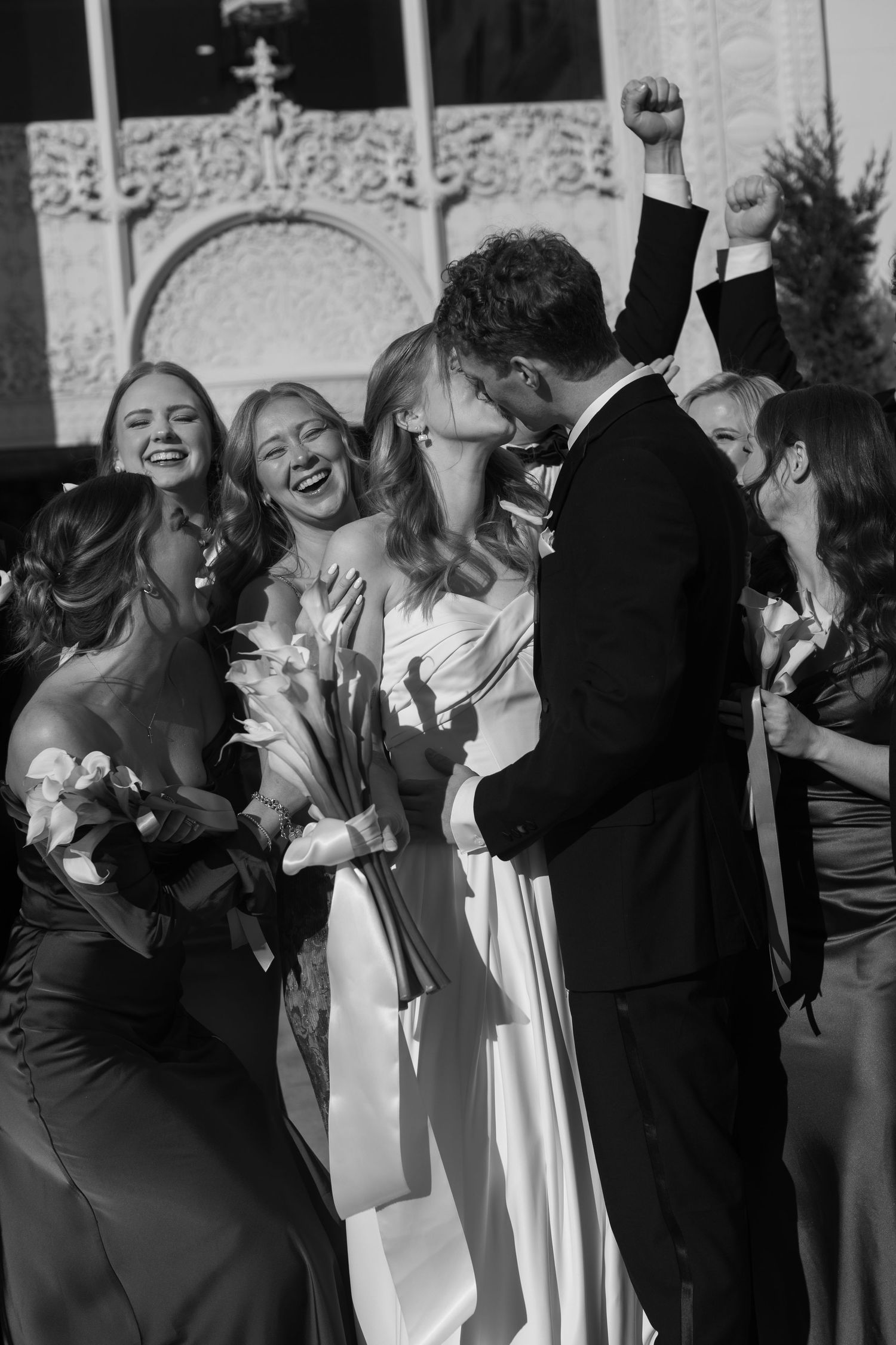 A joyful wedding party celebrates and poses together outside a building in black and white.