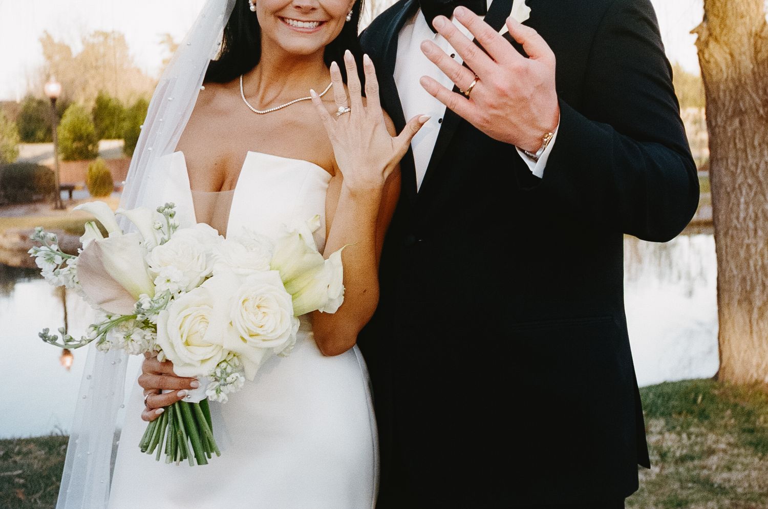 A couple in wedding attire poses outdoors holding a white floral bouquet.