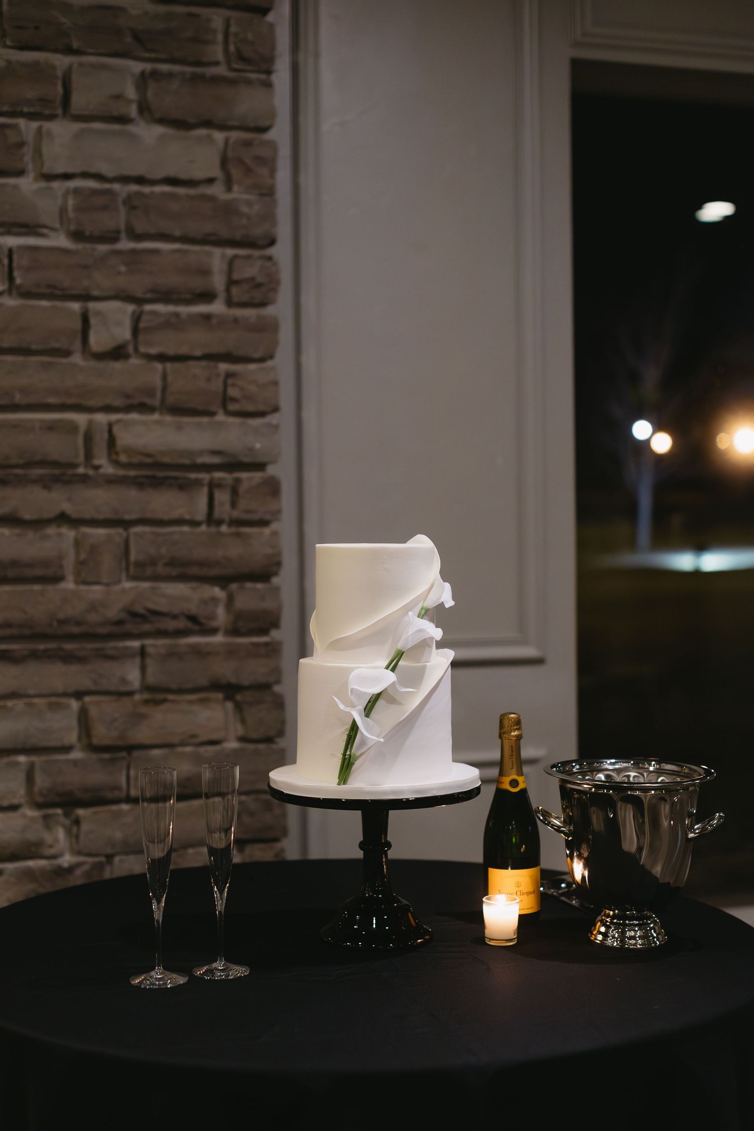 Elegant white wedding cake with champagne and glasses on black tablecloth against stone wall.