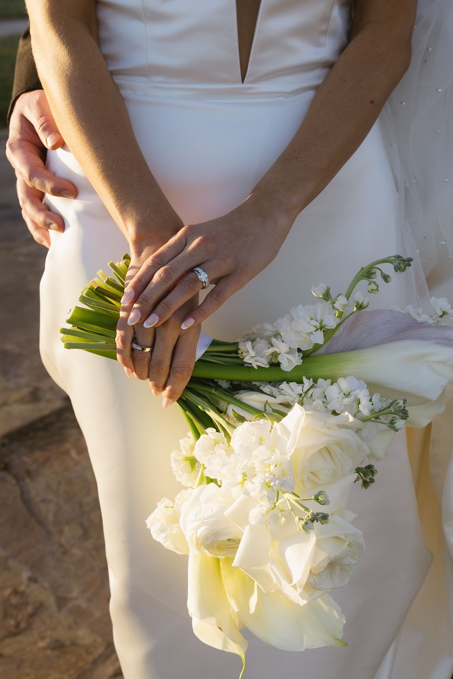 Hands holding white calla lily bouquet with engagement ring prominently displayed.