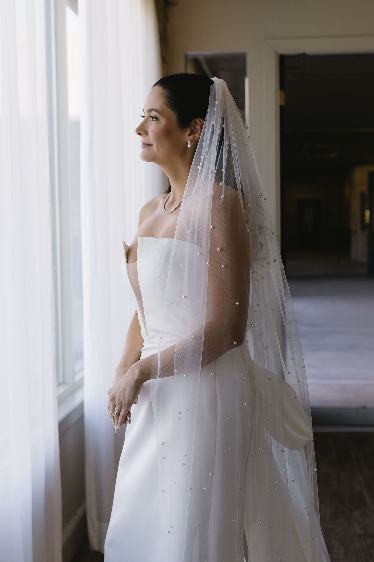 Bride wearing pearl-studded veil poses near sunlit windows in wedding venue.