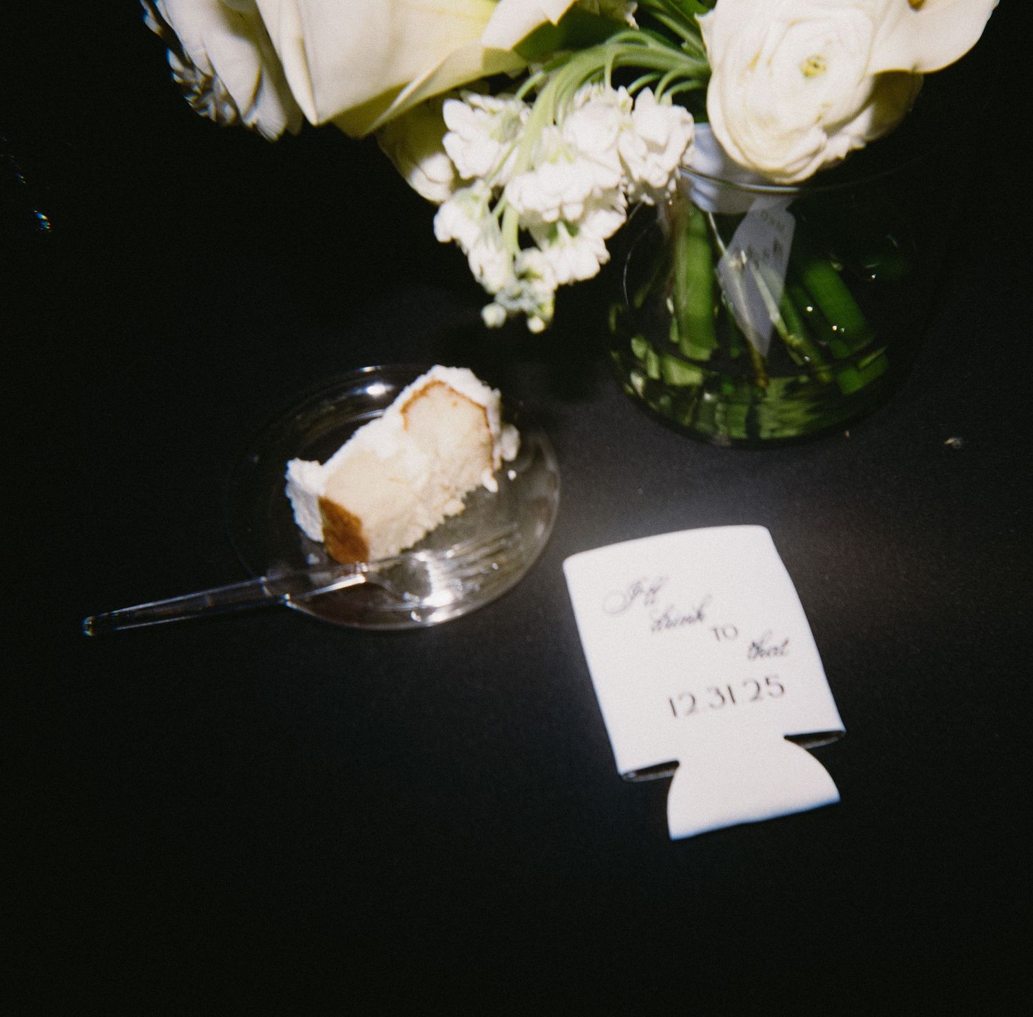 Wedding reception detail with white roses in a vase next to a drink koozie and cake slice.