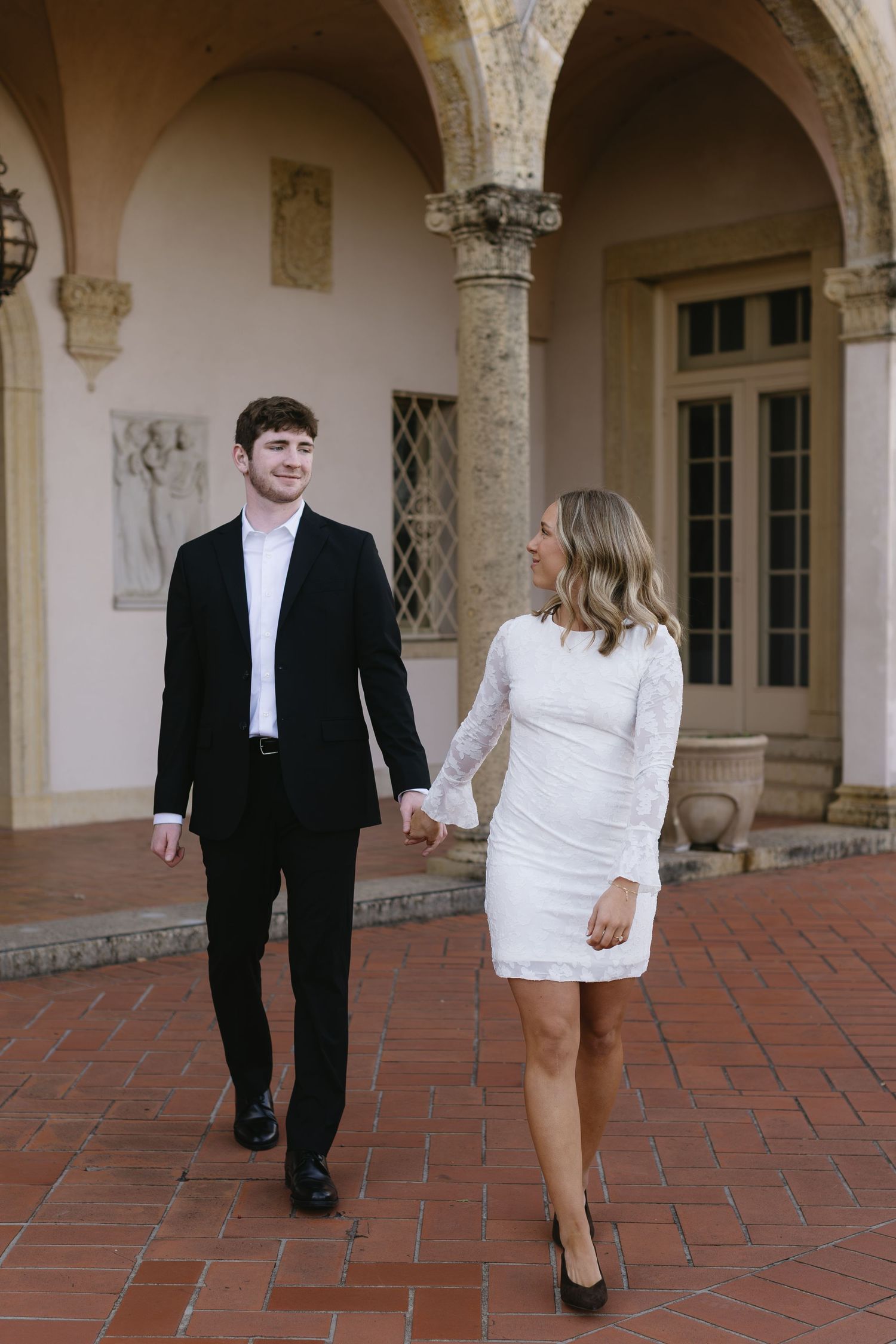 A couple in formal attire walks along a Spanish colonial style courtyard with arched columns.