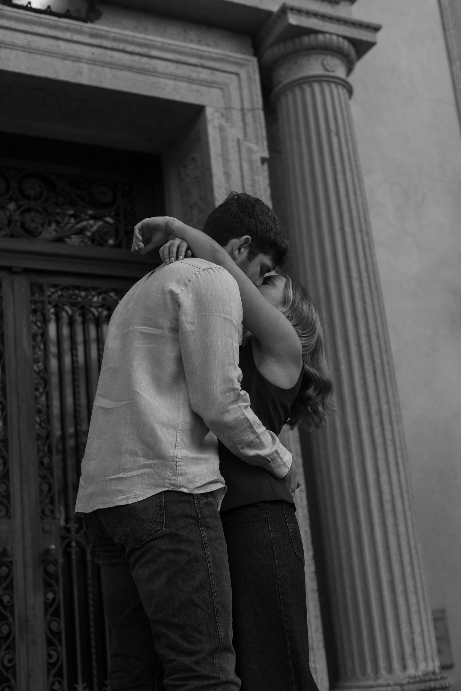 A black and white romantic photo of a couple kissing near a classical column.
