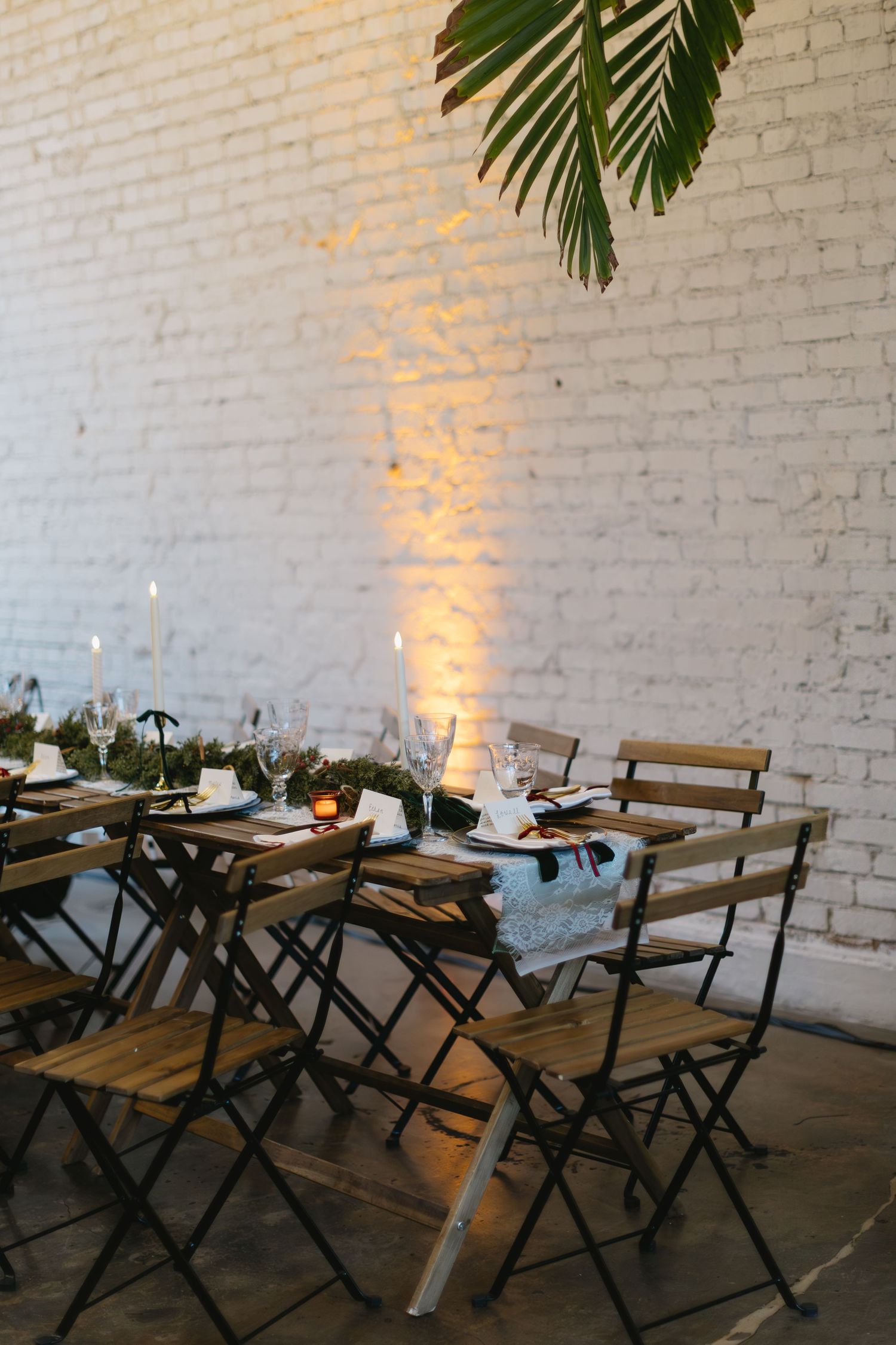 An elegant wedding reception table setting with palm leaves and warm lighting against a white brick wall.