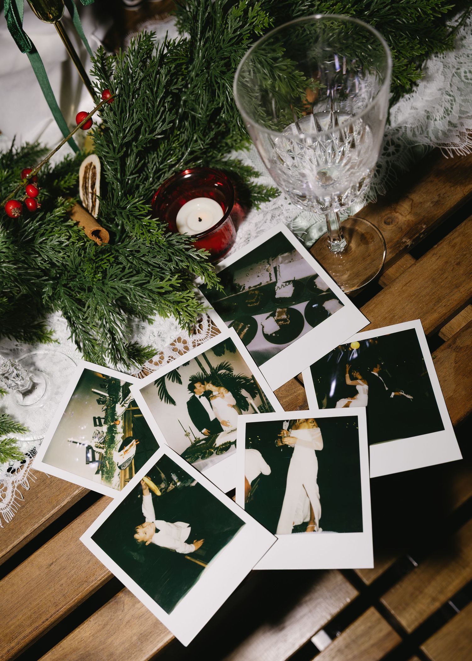 Holiday table setting with scattered Polaroid photos, evergreen branches, and wine glasses.