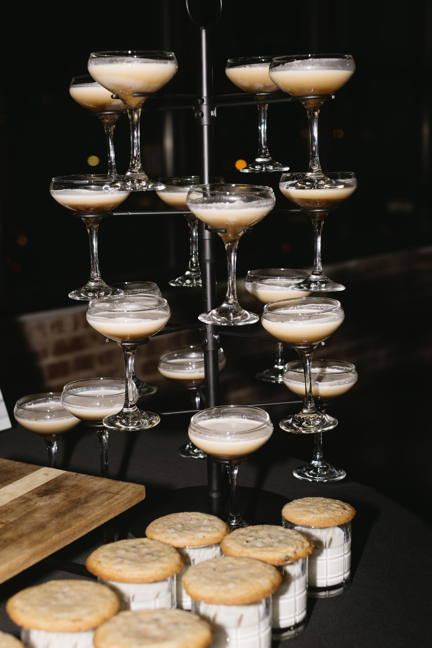 Rows of champagne cocktails in coupe glasses arranged on dark surface with cookies below.