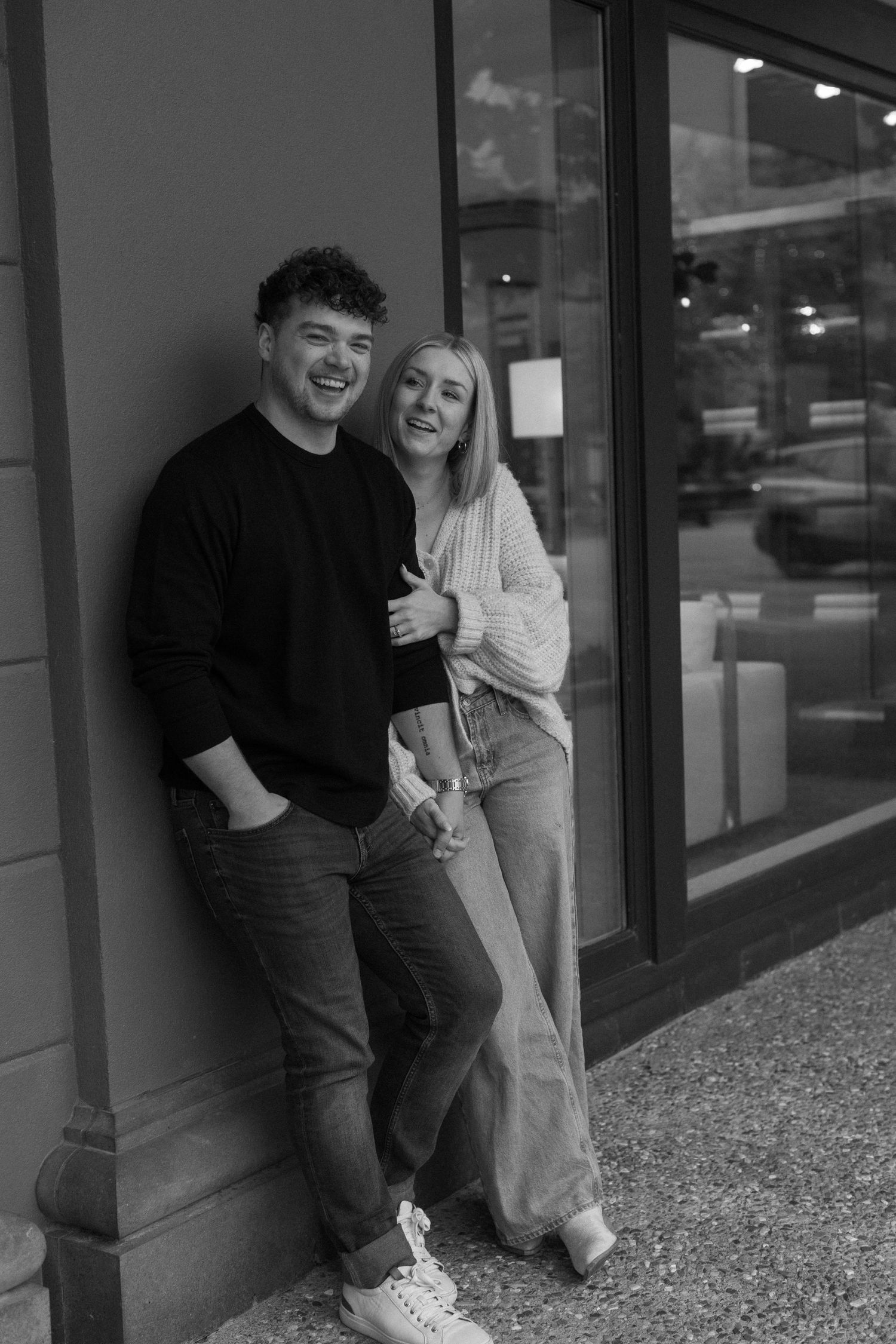 A black and white portrait of a couple sharing a happy moment outside a storefront.