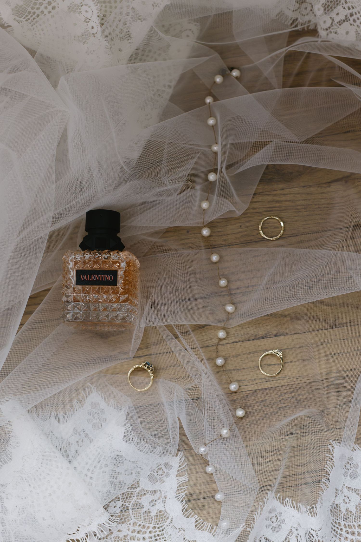 Wedding details including Valentino perfume bottle, delicate lace veil, and gold rings arranged on wooden surface.