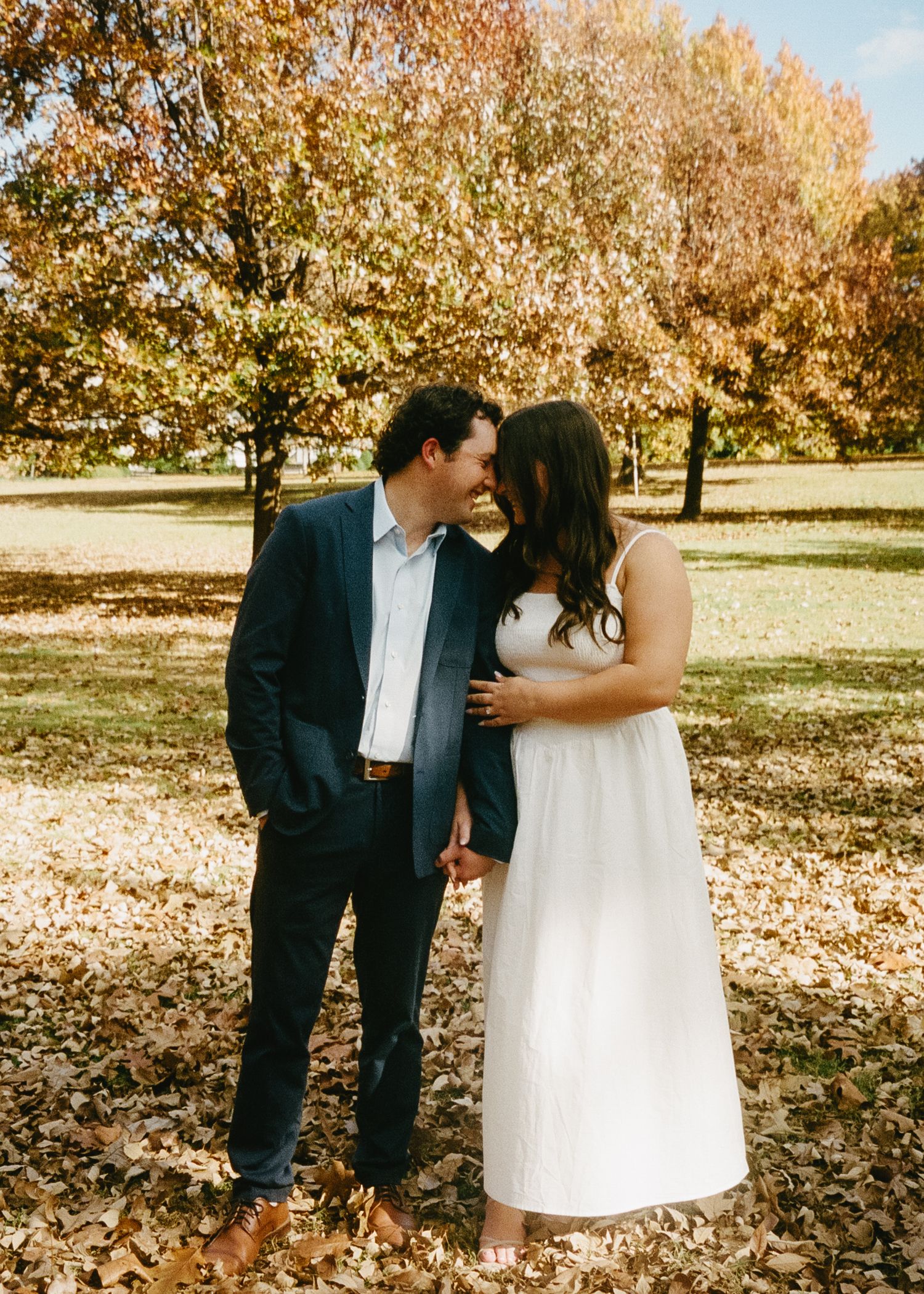A couple shares a romantic moment while walking through autumn leaves in a sunlit park on a fall day.
