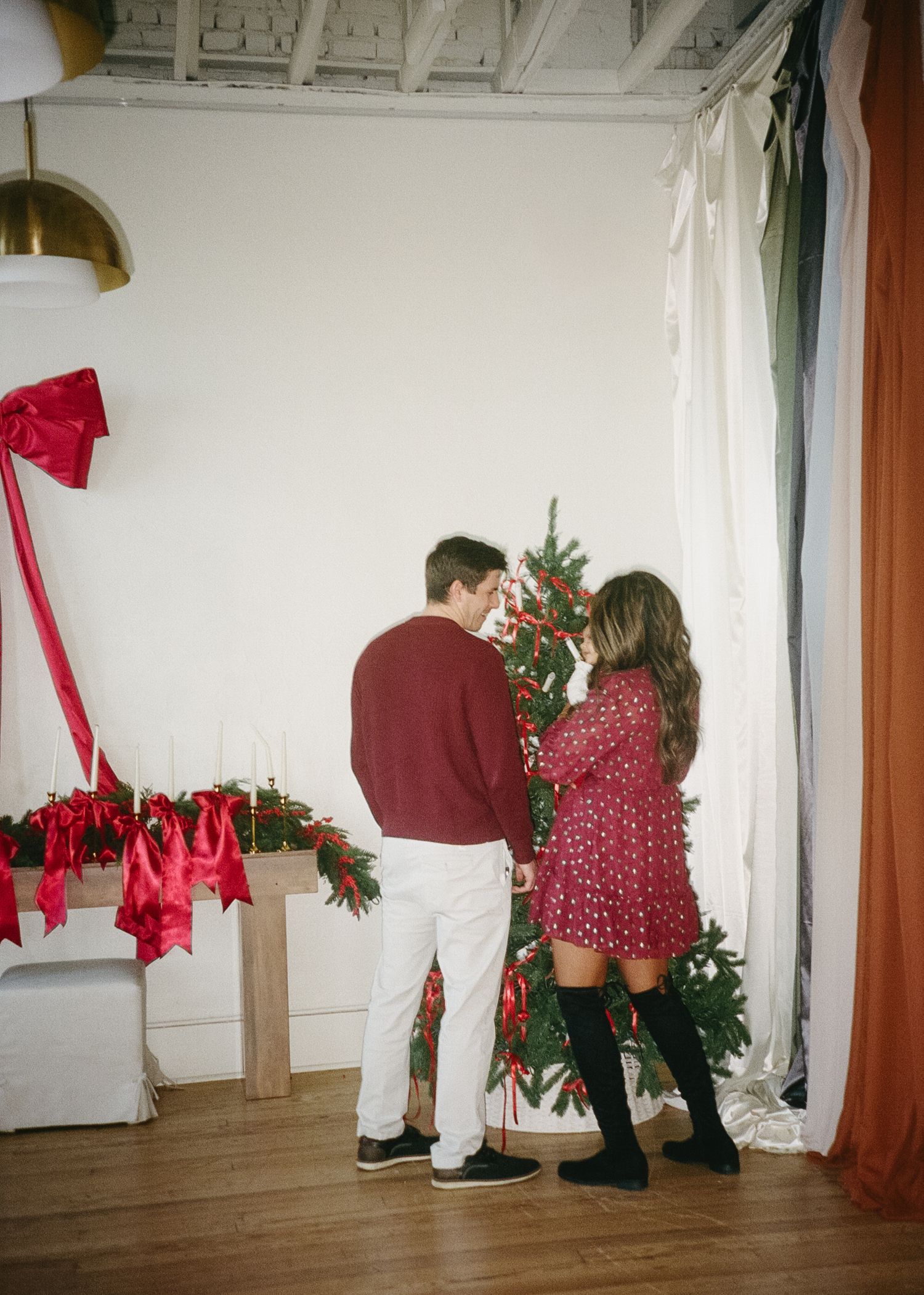 A couple decorates a Christmas tree together in a room with red holiday decor and white walls.