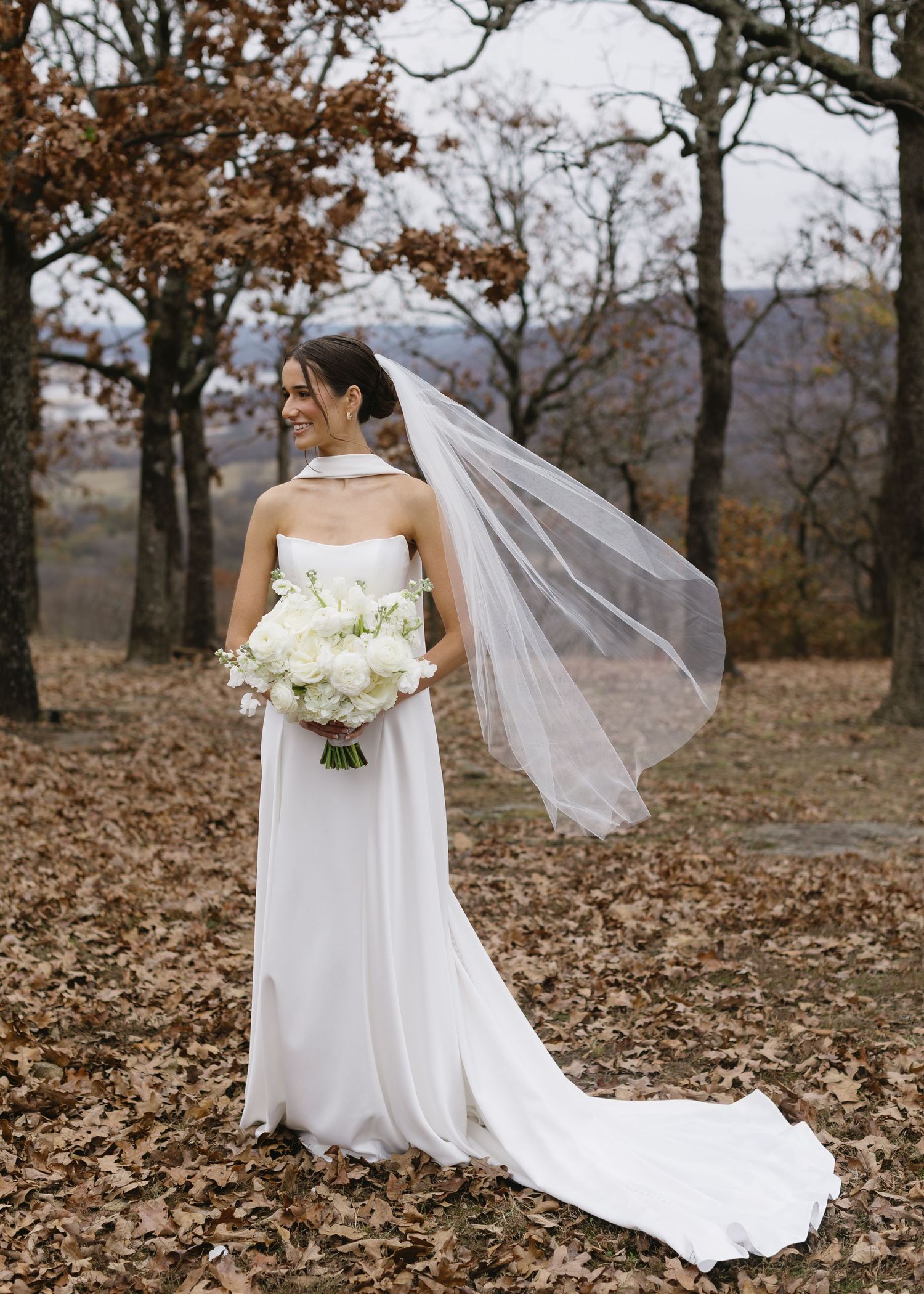 A bride in an elegant white dress holds a bouquet while standing among fall trees.