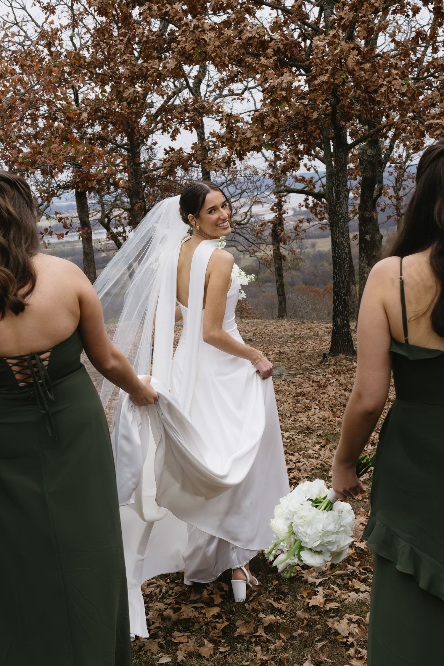 A bride in white walks with bridesmaids in dark dresses through a fall forest setting.