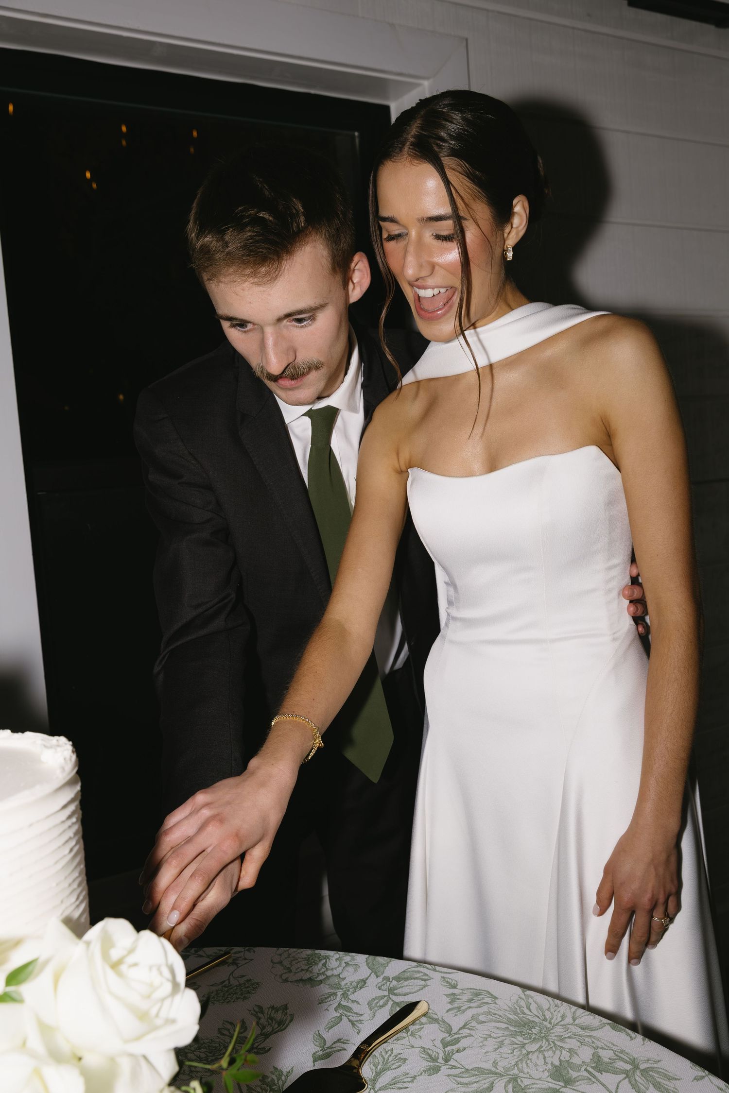 Newlyweds in formal attire cut their white wedding cake together during their reception celebration.