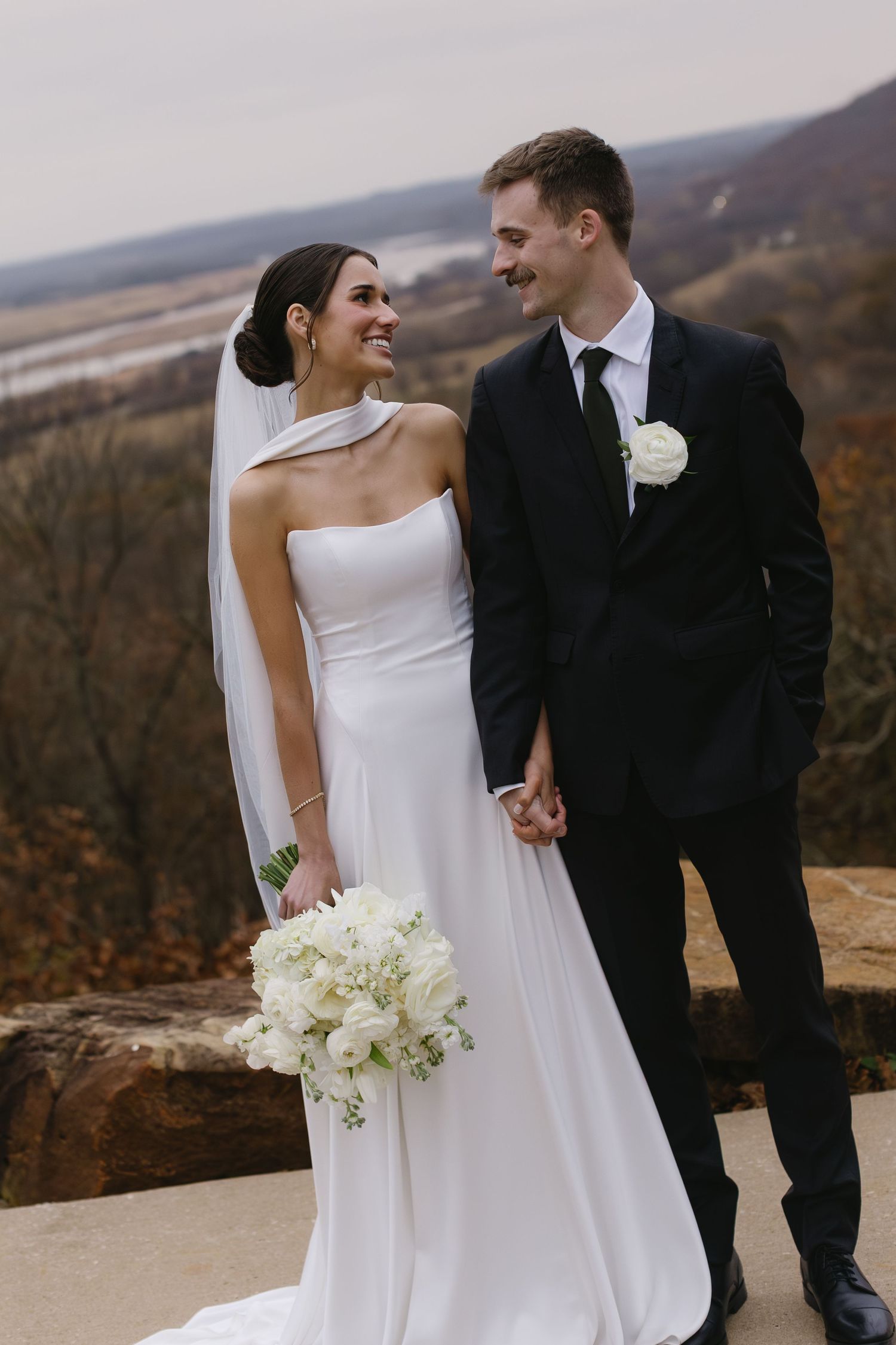 Joyful wedding couple poses on scenic overlook holding white floral bouquet.