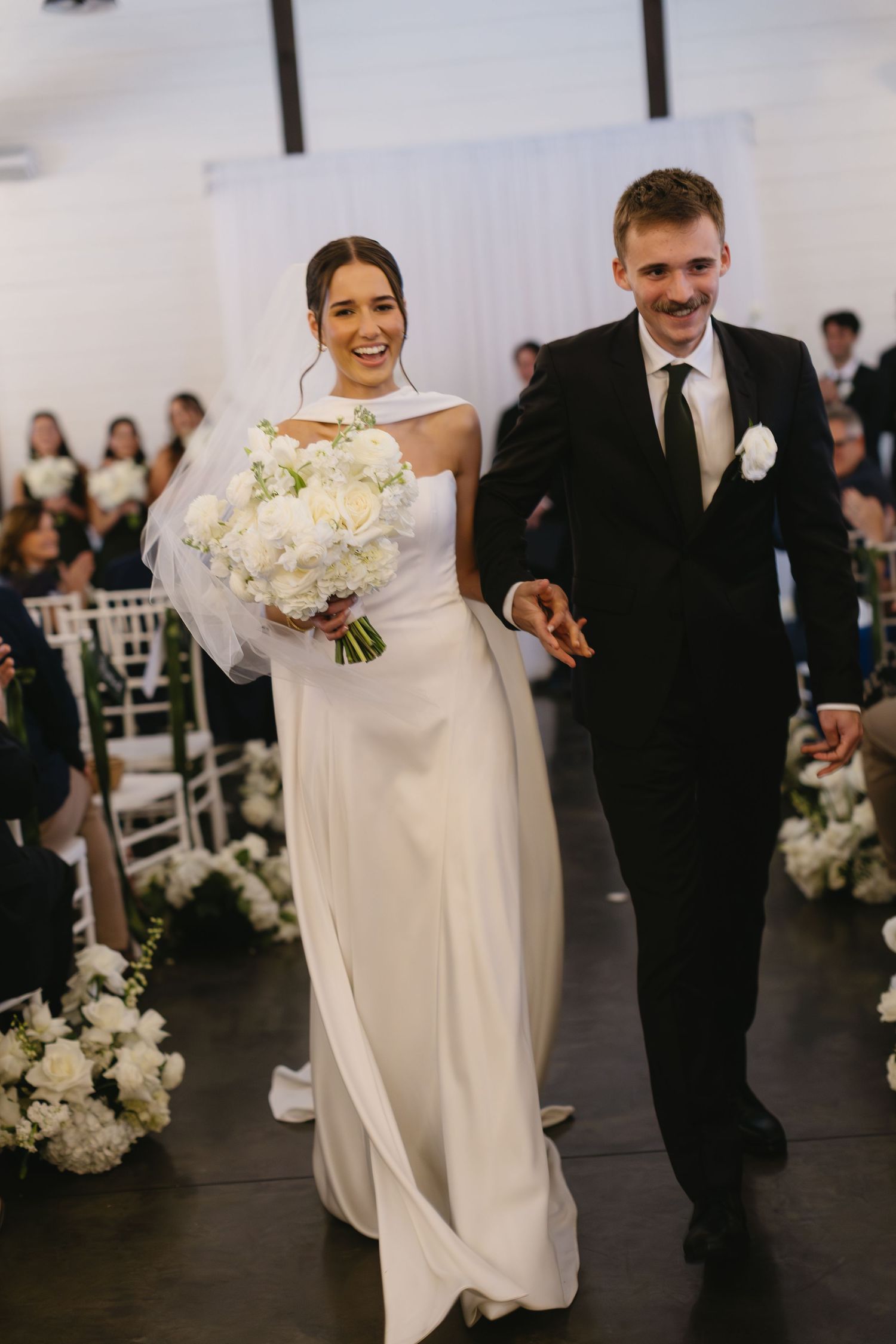 Wedding party members walk in formal procession through an elegant indoor venue with white decor.