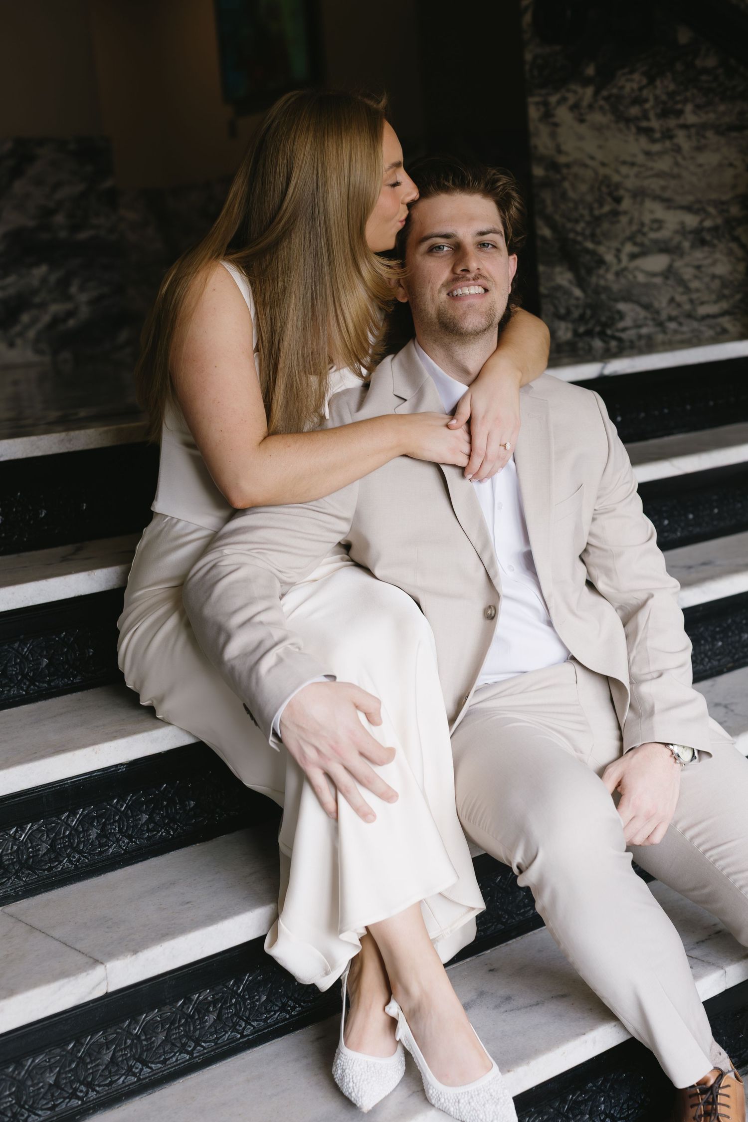 A couple in white attire shares playful moments while sitting on concrete steps.