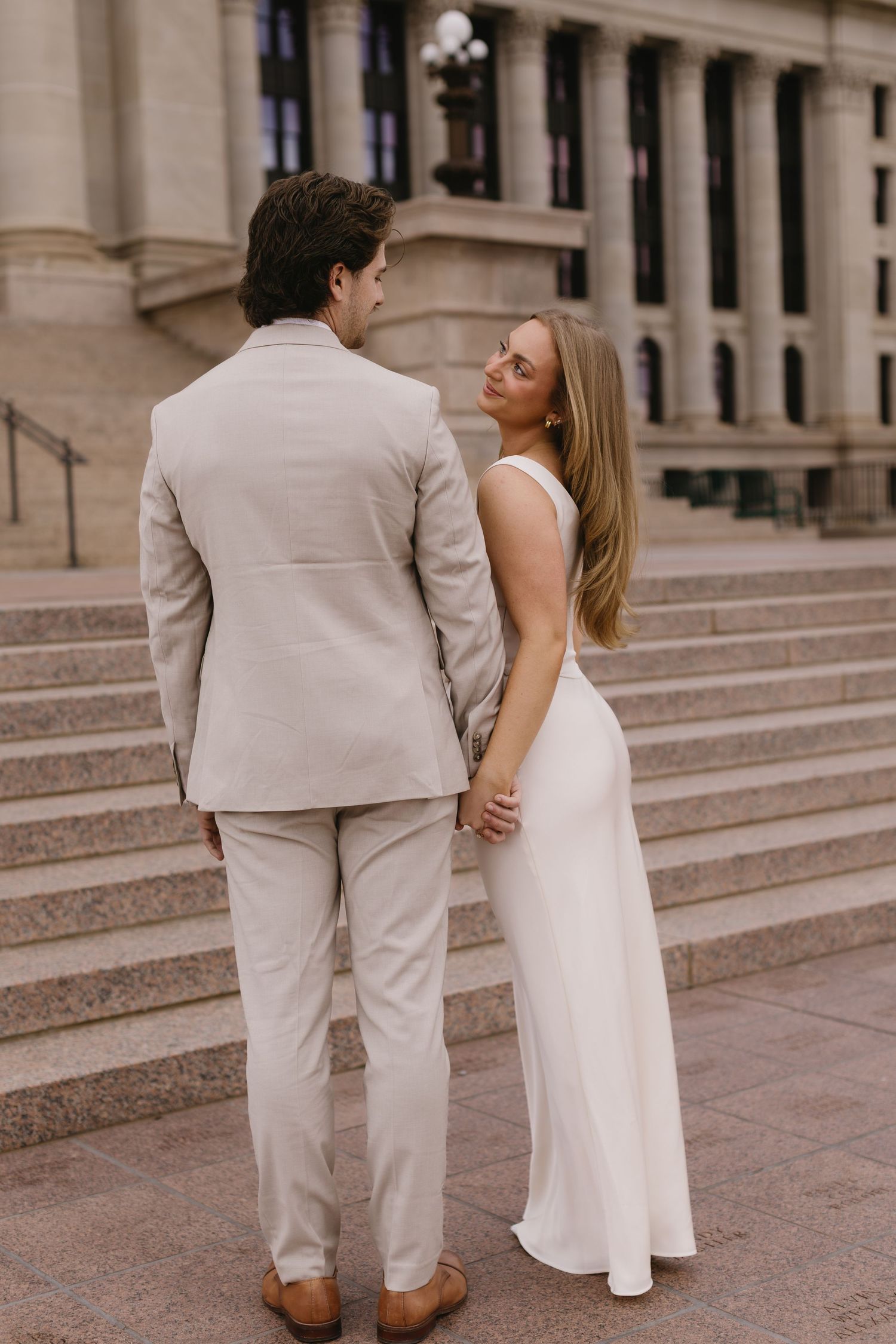 A couple in elegant beige and white attire pose romantically on grand stone steps of a historic building.