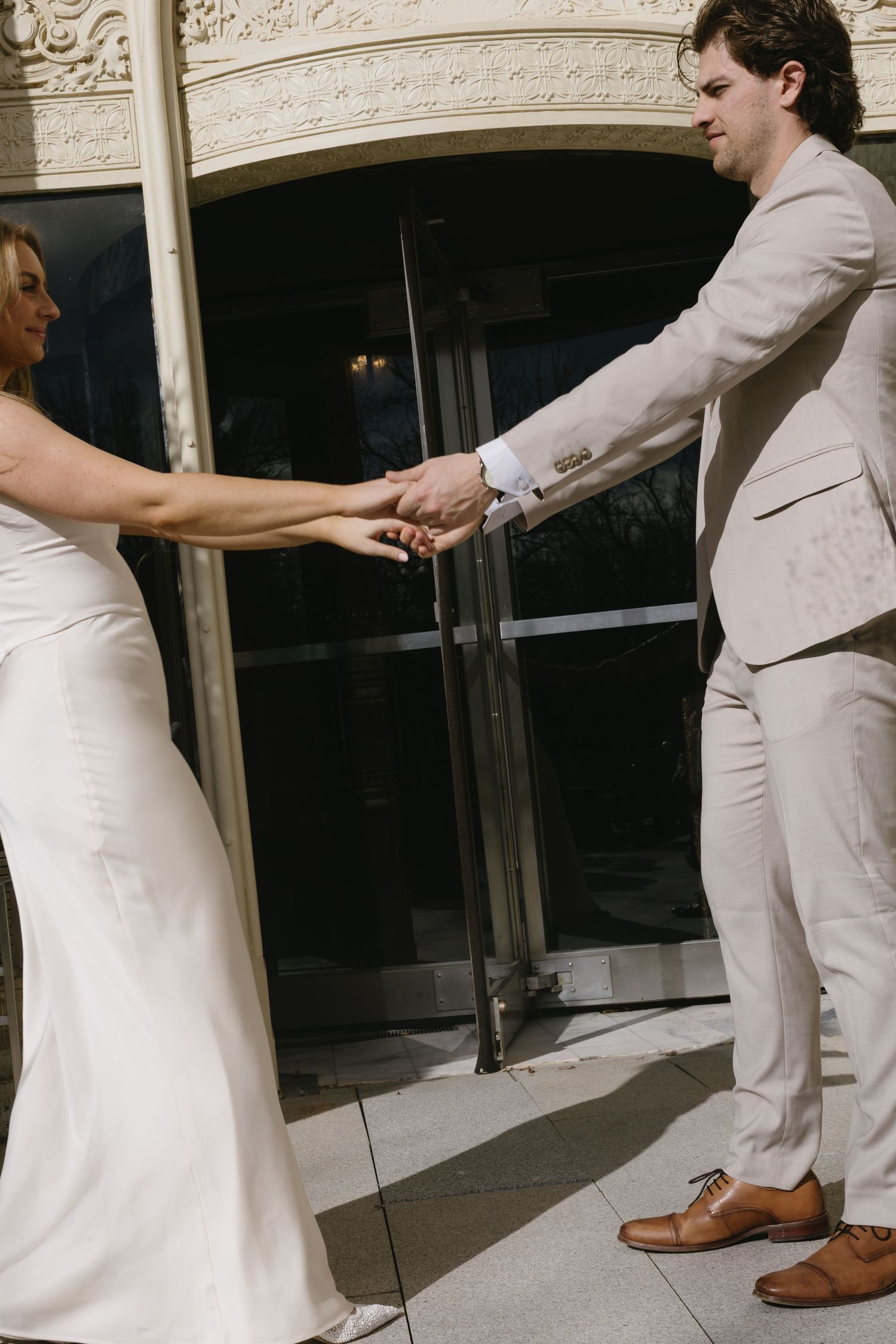 A playful moment between a couple in formal wear holding hands by glass doors.