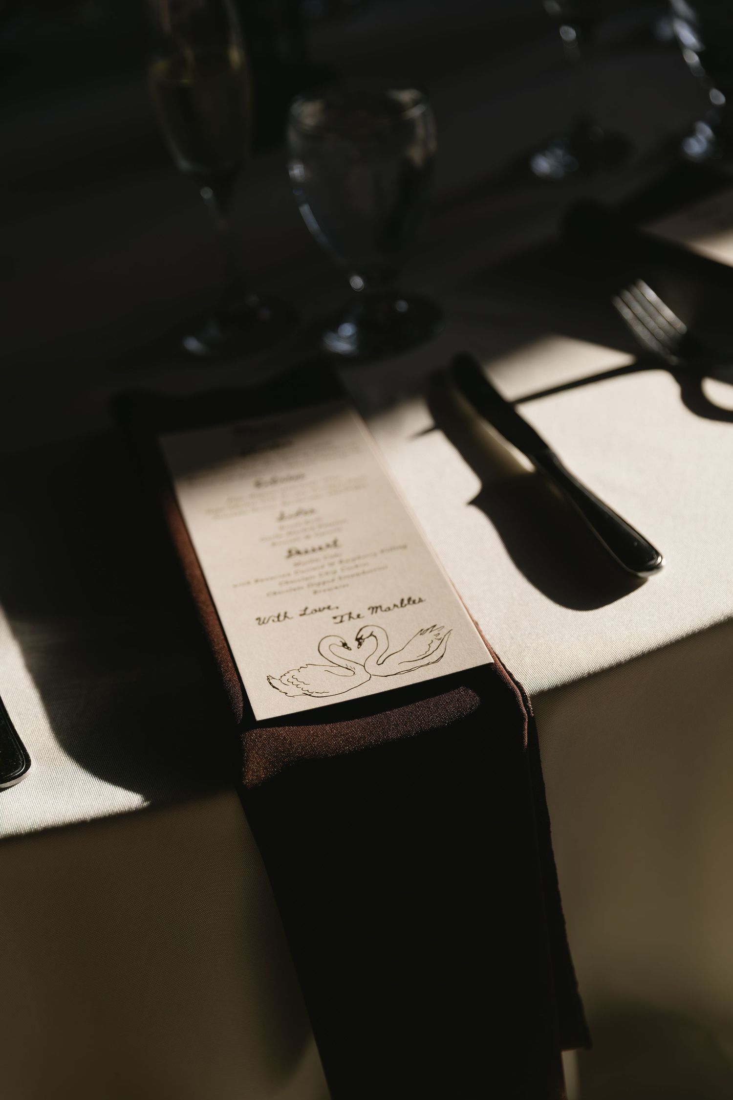 Close-up of a wedding menu card with a heart design in moody lighting with wine glasses casting shadows.