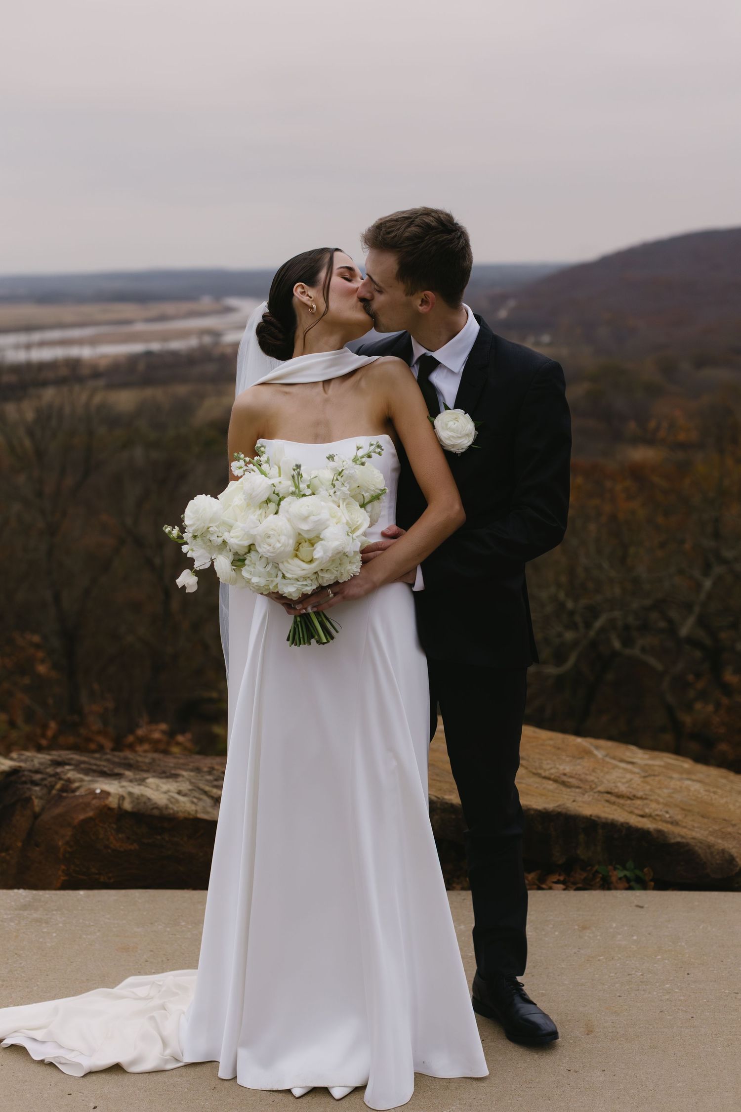 A romantic wedding couple shares a kiss on a scenic mountain overlook with autumn foliage in the background.