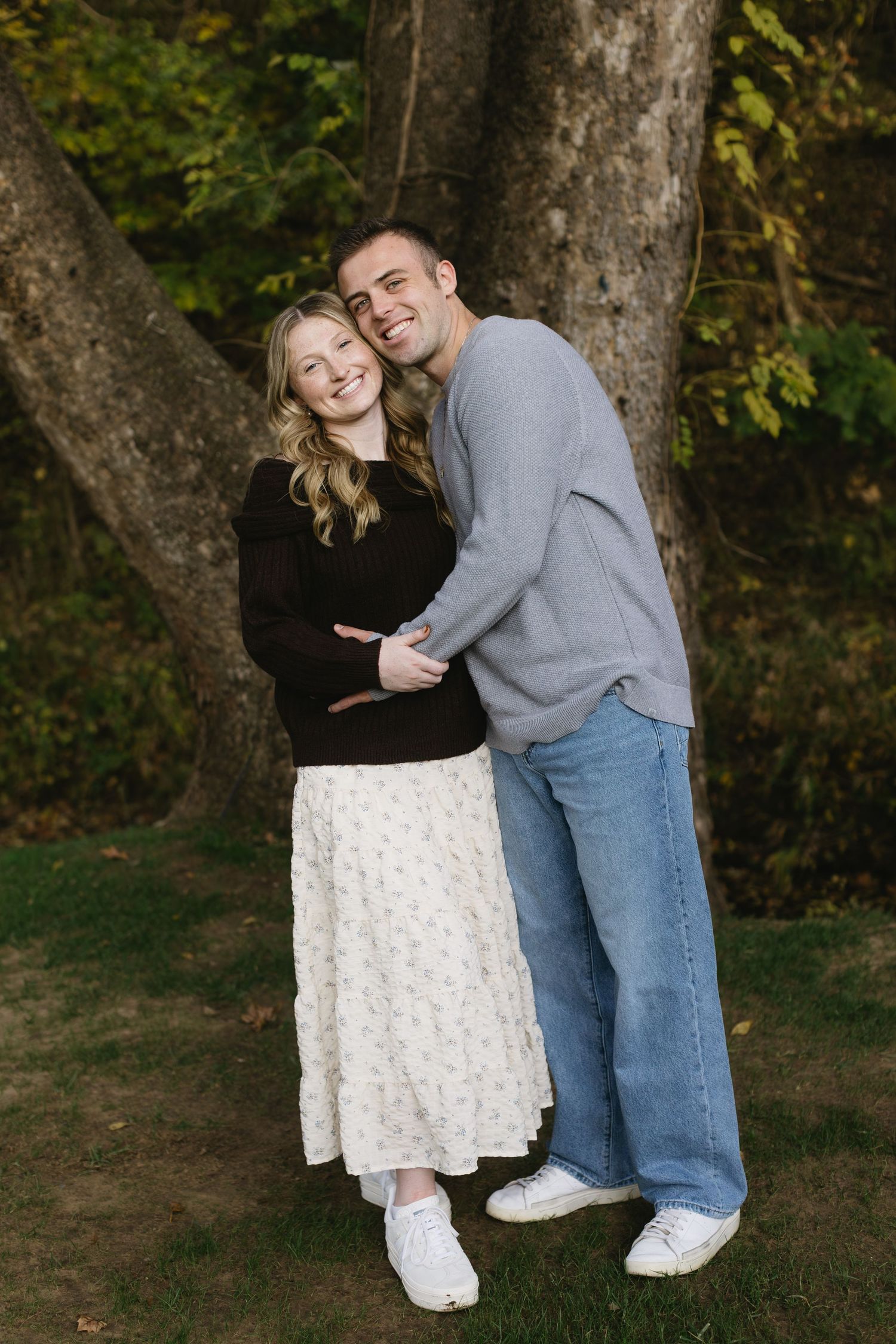 A playful couple poses together against tree trunks in a park setting.