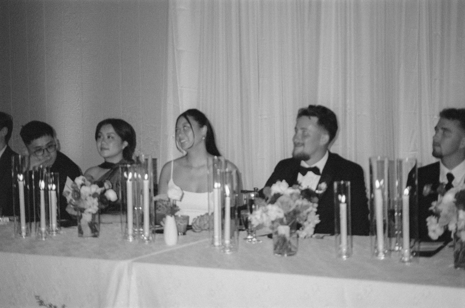 Wedding party seated at head table with tall candles and floral arrangements.
