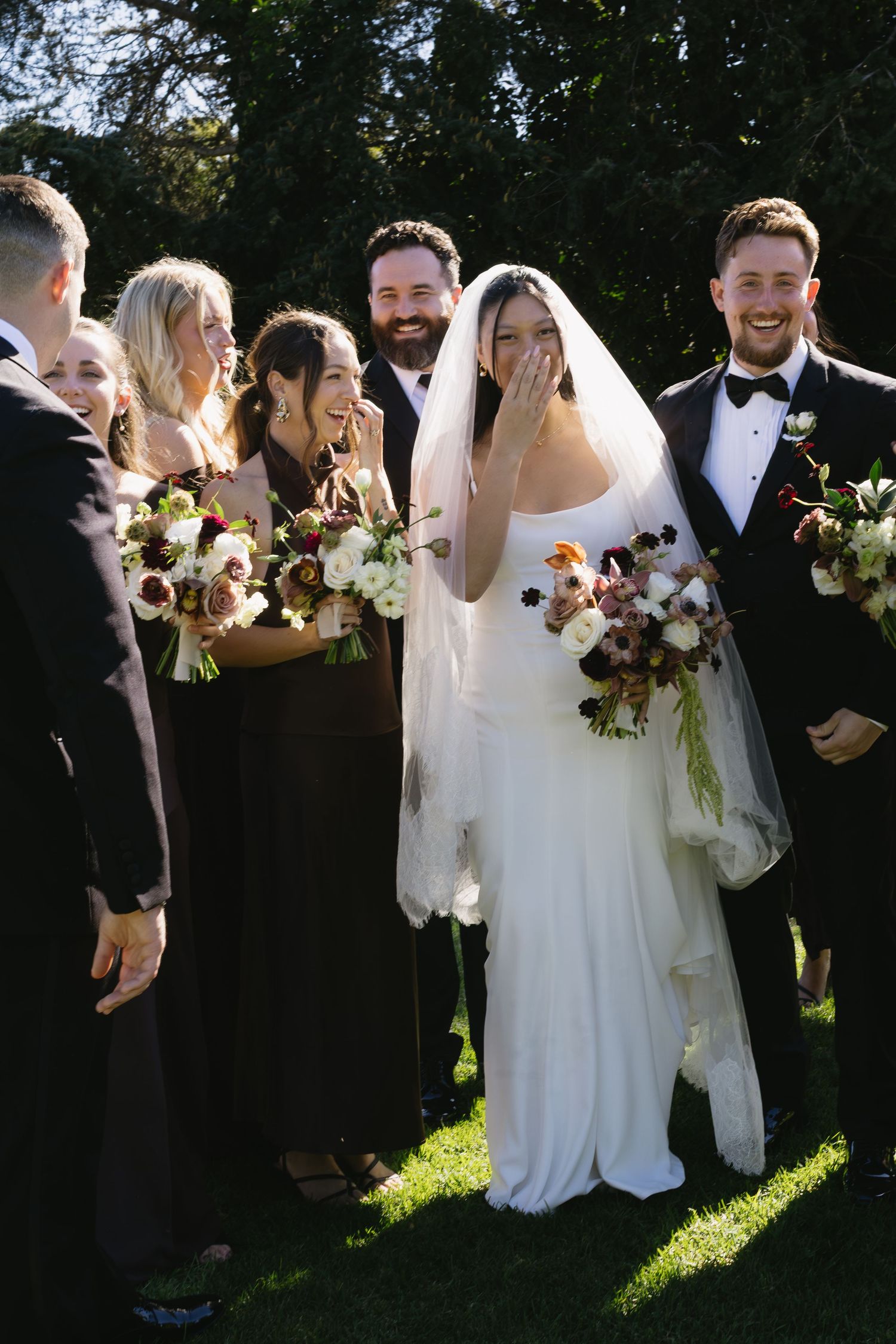 Wedding party laughs together during outdoor photo session with sunlight filtering through trees.