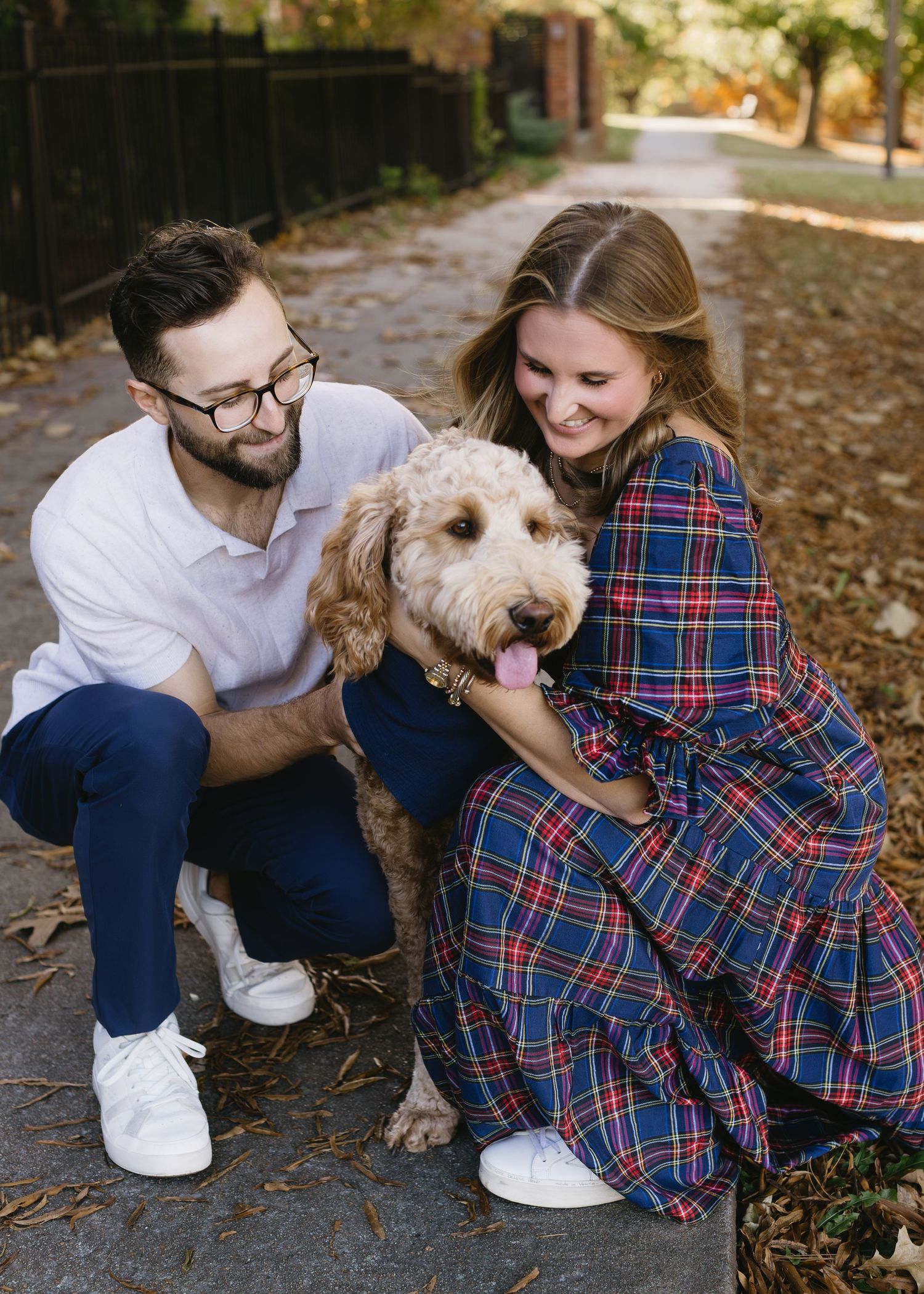 A couple sits outdoors with their fluffy goldendoodle dog on an autumn day with fallen leaves.